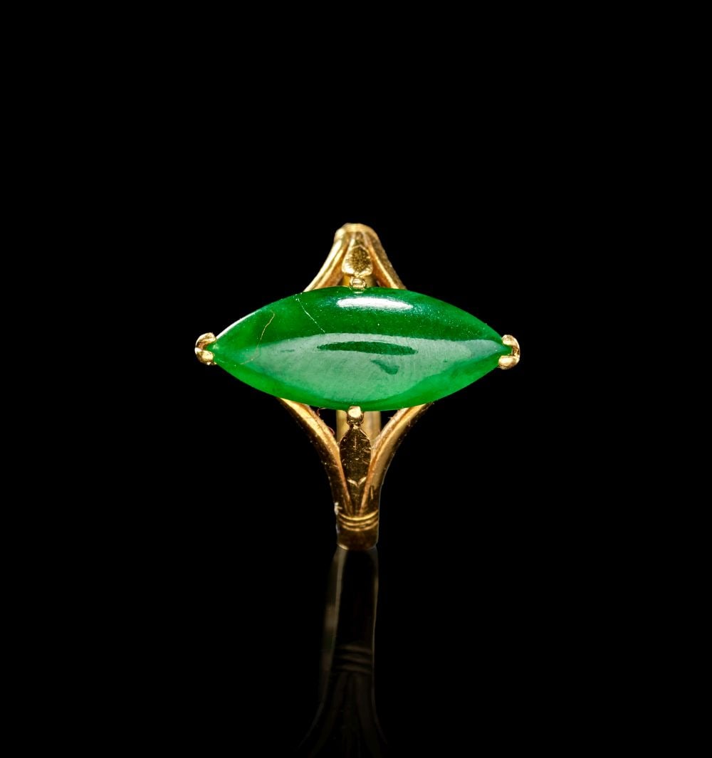 A CHINESE JADE & GOLD RING, REPUBLIC PERIOD: A CHINESE JADE & GOLD RING, REPUBLIC PERIOD The ring is set with an elongated marquise-shaped green jade cabochon, mounted horizontally within a gold claw setting. The stone has a polished, glassy