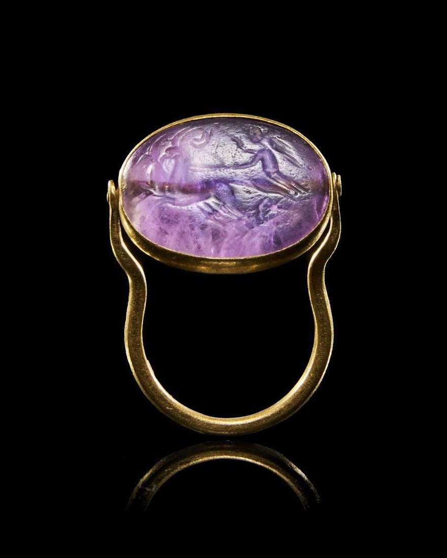 A ROMAN AMETHYST RING STONE DEPICTING CHARIOT RACING SCENE Circa 2nd Century A.D.: A ROMAN AMETHYST RING STONE DEPICTING CHARIOT RACING SCENECirca 2nd Century A.D.An oval amethyst intaglio set within a smooth gold ring, the stone a rich translucent purple with natural internal veini