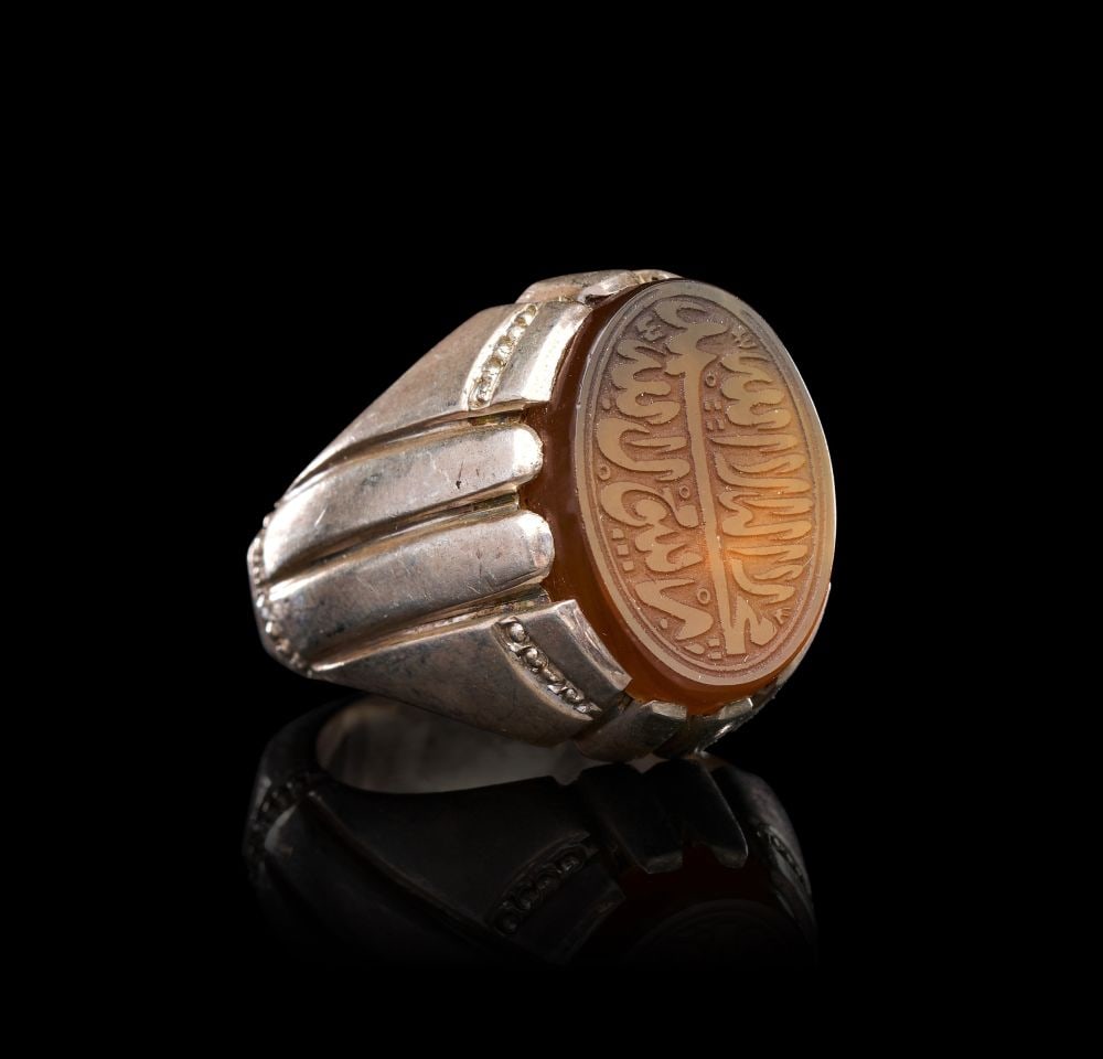 A CALLIGRAPHIC INSCRIBED AGATE RING ON SILVER, 19TH CENTURY, PERSIA: A CALLIGRAPHIC INSCRIBED AGATE RING ON SILVER, PERSIA, 19TH CENTURY The oval bezel set with a honey-toned agate of even translucency, the stone intaglio-carved in reverse with a multi-line Arabic insc