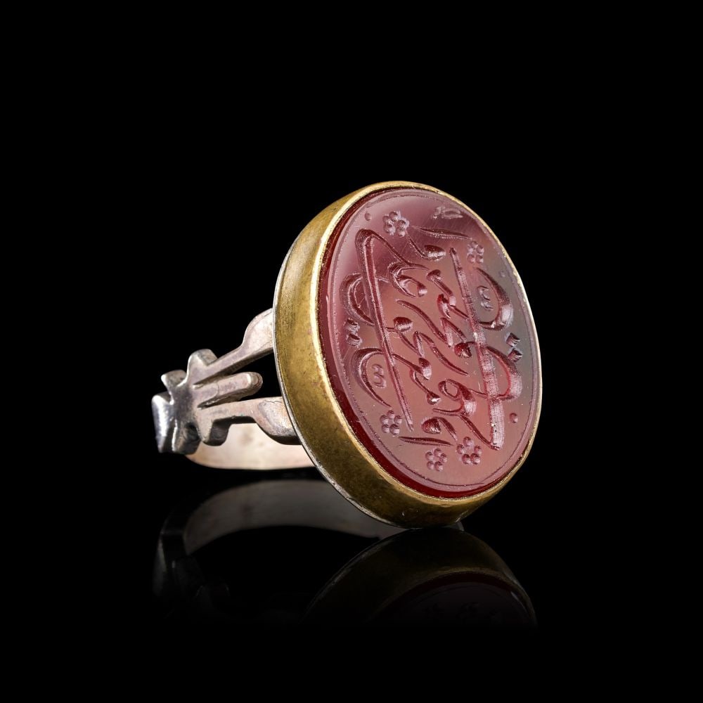 A CALLIGRAPHIC INSCRIBED AGATE RING ON SILVER, 19TH CENTURY, PERSIA: A CALLIGRAPHIC INSCRIBED AGATE RING ON SILVER, 19TH CENTURY, PERSIA The ring is set with an oval agate of warm reddish tone, held within a plain gilt-metal collet raised on a silver hoop. The shoulder
