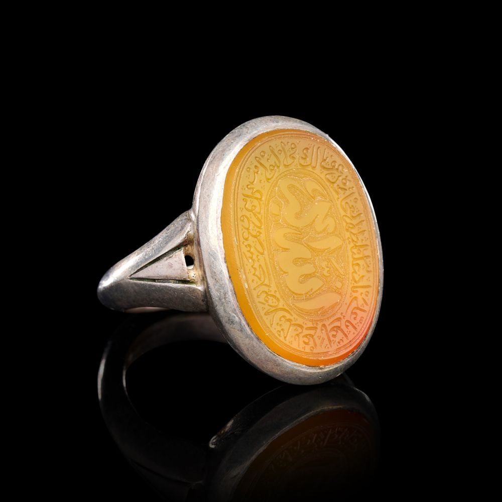 A CALLIGRAPHIC INSCRIBED AGATE RING ON SILVER, 19TH CENTURY, PERSIA: A CALLIGRAPHIC INSCRIBED AGATE RING ON SILVER, PERSIA, 19TH CENTURY The ring of oval bezel-form, the broad silver hoop rising from a flat shank through openwork triangular shoulders to a deep collet s