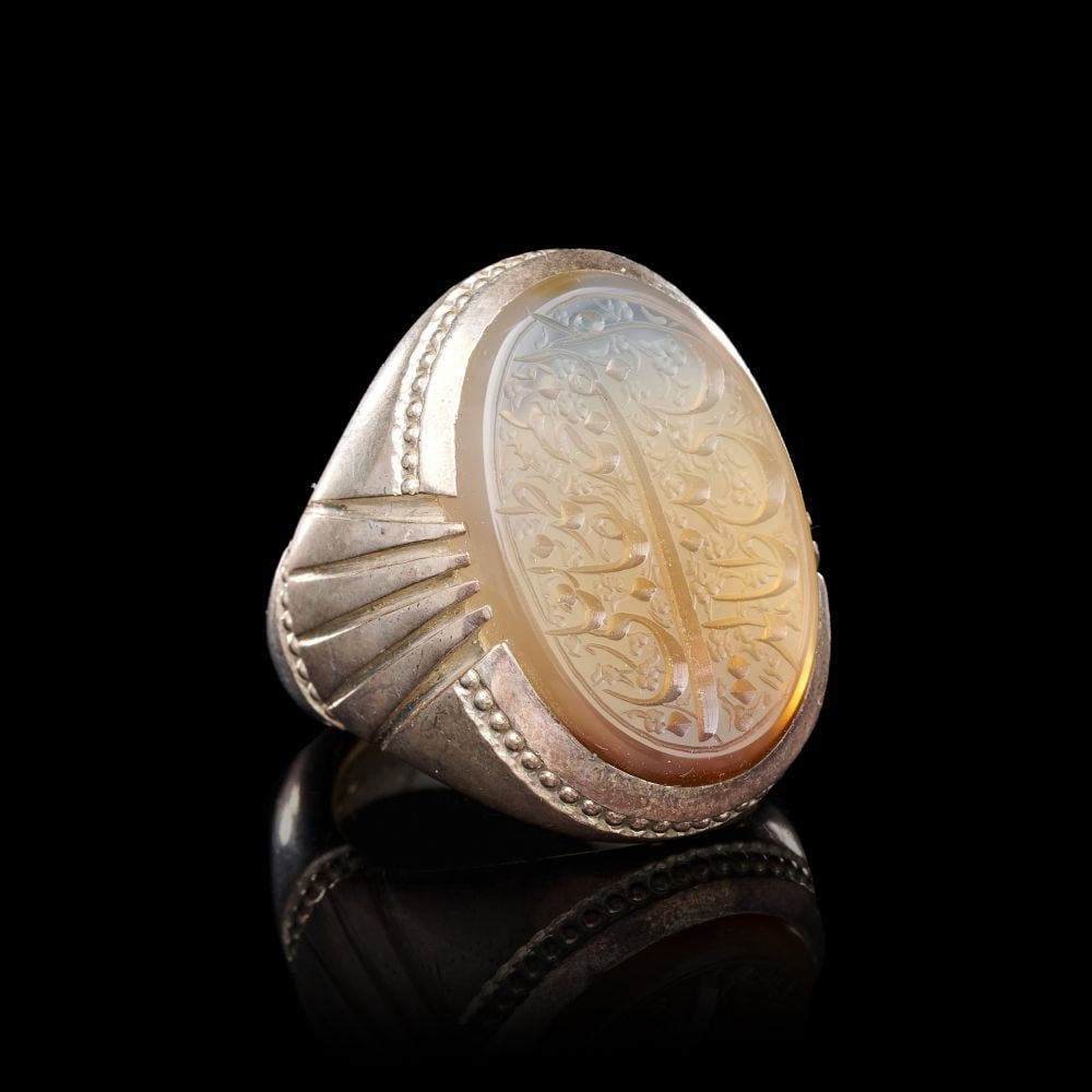 A CALLIGRAPHIC INSCRIBED AGATE RING ON SILVER, 19TH CENTURY, PERSIA: A CALLIGRAPHIC INSCRIBED AGATE RING ON SILVER, PERSIA, 19TH CENTURY The ring of substantial form, set with a large oval agate bezel of pale honey tone shading to translucent grey at the upper edge, th