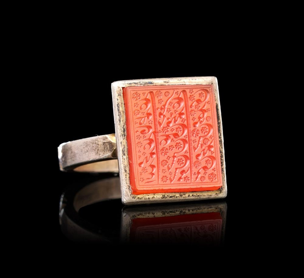 A CALLIGRAPHIC INSCRIBED AGATE RING ON SILVER, 19TH CENTURY, PERSIA: A CALLIGRAPHIC INSCRIBED AGATE RING ON SILVER, PERSIA, 19TH CENTURY Of substantial form, the rectangular bezel set with a deeply carved orange-red agate plaque held within a plain silver collet, the s
