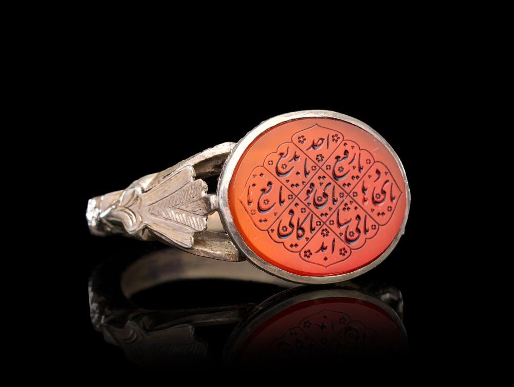 A CALLIGRAPHIC INSCRIBED AGATE RING ON SILVER, 19TH CENTURY, PERSIA: A CALLIGRAPHIC INSCRIBED AGATE RING ON SILVER, PERSIA, 19TH CENTURY The ring of substantial form, the silver hoop rising from a slender shank to broad openwork shoulders chased and engraved with styli