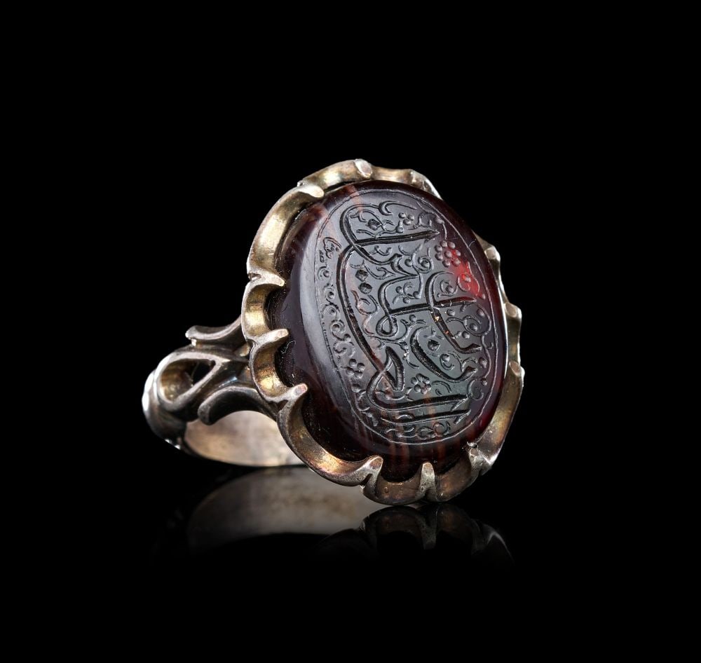 A CALLIGRAPHIC INSCRIBED AGATE RING ON SILVER, 19TH CENTURY, PERSIA: A CALLIGRAPHIC INSCRIBED AGATE RING ON SILVER, PERSIA, 19TH CENTURY Of oval form, the bezel set with a deep red-brown banded agate cabochon held within a silver mount of scalloped, foliate claws, the 