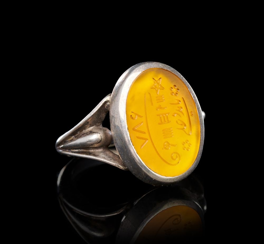 AN UNUSUAL CALLIGRAPHIC INSCRIBED AGATE RING ON SILVER, 19TH CENTURY, PERSIA: AN UNUSUAL CALLIGRAPHIC INSCRIBED AGATE RING ON SILVER, 19TH CENTURY, PERSIA The ring of substantial form, the silver hoop rising from a tapered shank with openwork shoulders that flare and meet the o