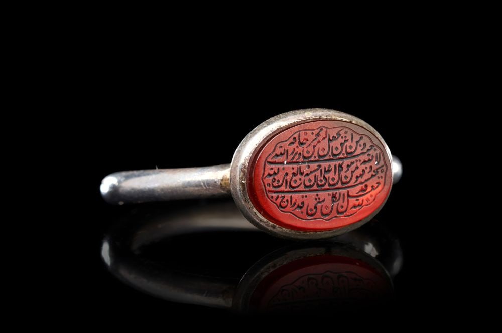 A CALLIGRAPHIC INSCRIBED AGATE RING ON SILVER, 19TH CENTURY, PERSIA: A CALLIGRAPHIC INSCRIBED AGATE RING ON SILVER, PERSIA, 19TH CENTURY Of typical form, the silver hoop of plain circular section rising to a broad oval bezel set with a flat carnelian-coloured agate int