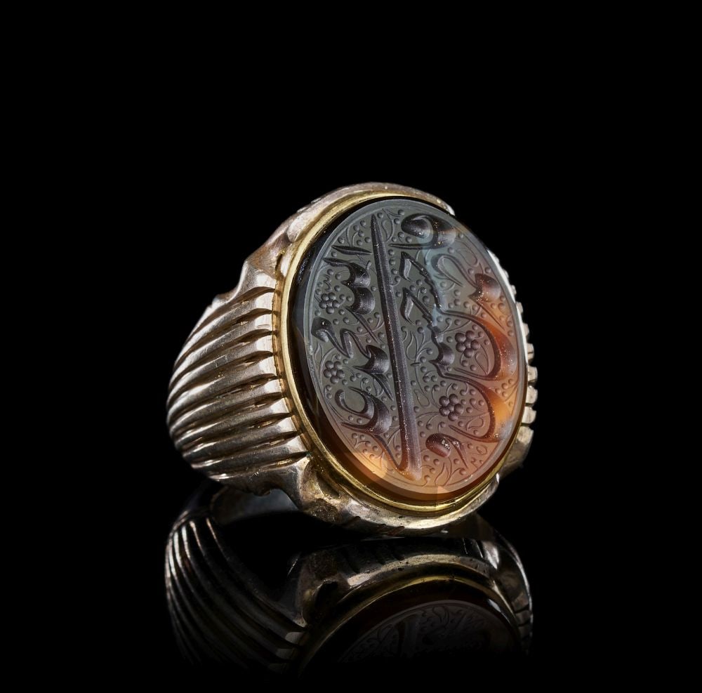 A CALLIGRAPHIC INSCRIBED AGATE RING ON SILVER, 19TH CENTURY, PERSIA: A CALLIGRAPHIC INSCRIBED AGATE RING, PERSIA, 19TH CENTURY Of substantial form, the oval agate bezel set within a plain gilt-metal collet and raised on a heavy silver hoop with deeply ribbed shoulders 