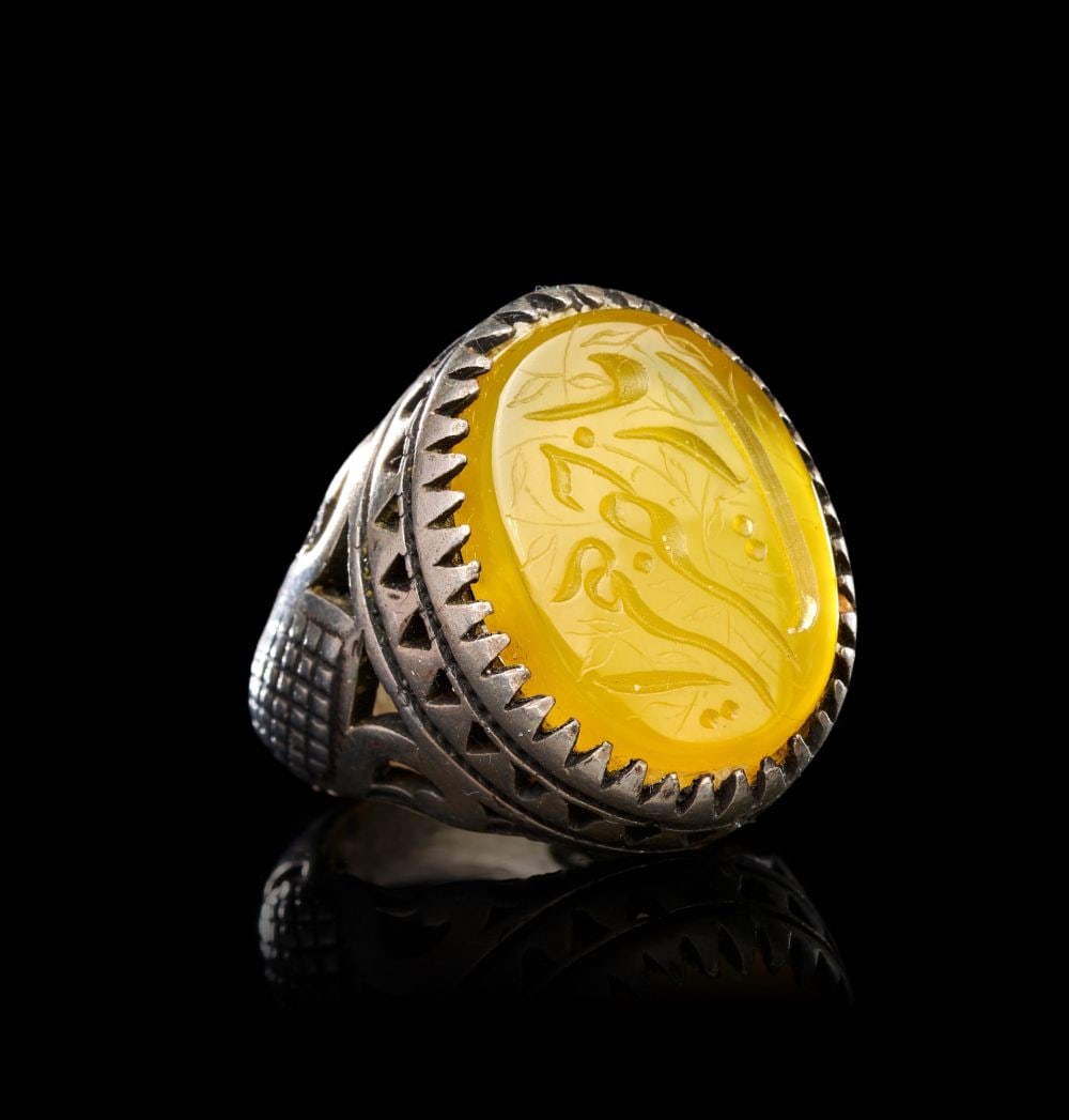 A CALLIGRAPHIC INSCRIBED AGATE RING ON SILVER, 19TH CENTURY, PERSIA: A CALLIGRAPHIC INSCRIBED AGATE RING ON SILVER, PERSIA, 19TH CENTURY Of oval bezel form, the ring set with a large translucent yellow agate cabochon engraved in intaglio with a flowing nasta'liq inscri