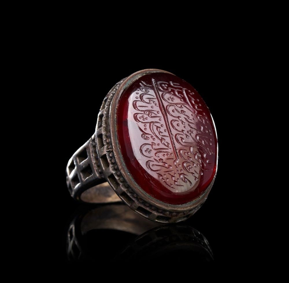 A CALLIGRAPHIC INSCRIBED AGATE RING ON SILVER, 19TH CENTURY, PERSIA: A CALLIGRAPHIC INSCRIBED AGATE RING ON SILVER, PERSIA, 19TH CENTURY Of substantial form, the ring set with a large oval cabochon of deep red agate held within a high silver collet, the bezel framed by
