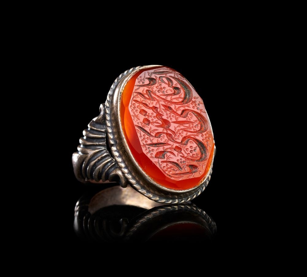 A CALLIGRAPHIC INSCRIBED AGATE RING ON SILVER, 19TH CENTURY, PERSIA: A QAJAR CALLIGRAPHIC CARNELIAN-INTAGLIO SILVER RING, PERSIA, 19TH CENTURY The ring of substantial form, the shoulders modelled with bold fluted and reeded volutes flanking a deep oval bezel, the stone