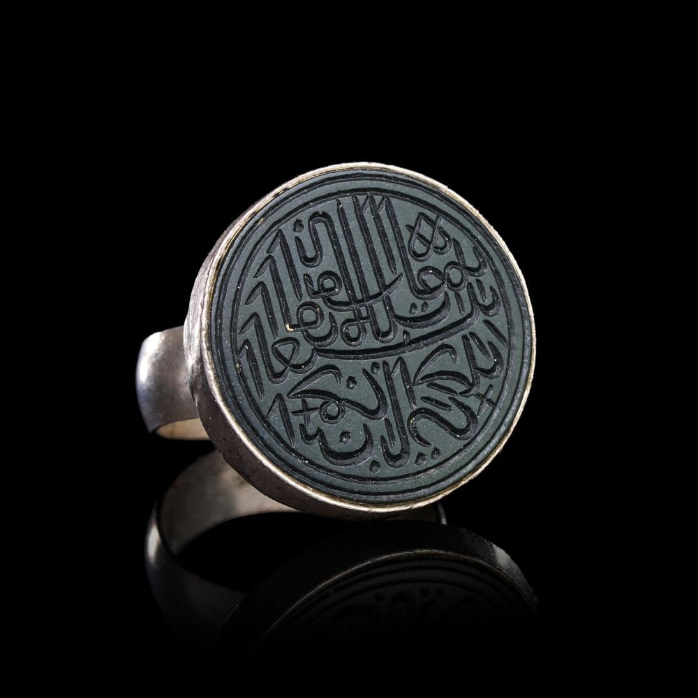 A CALLIGRAPHIC INSCRIBED JADE RING ON SILVER, 18TH CENTURY, PERSIA: A CALLIGRAPHIC INSCRIBED JADE RING ON SILVER, PERSIA, 18TH CENTURY The ring of substantial form, the broad silver shank rising to a circular bezel set with a disc of dark green jade, the stone framed 