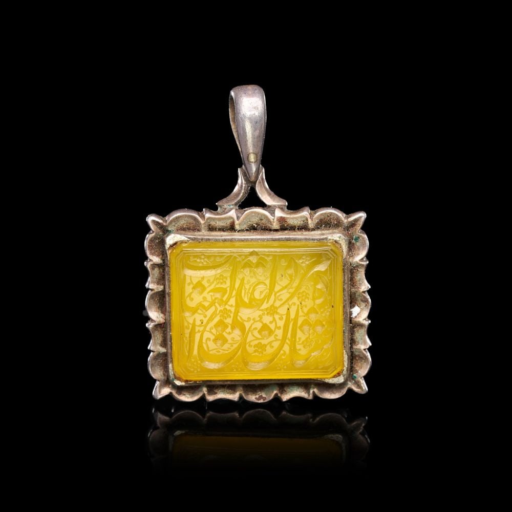 A CALLIGRAPHIC INSCRIBED AGATE AMULET ON SILVER, 19TH CENTURY, PERSIA: A CALLIGRAPHIC INSCRIBED AGATE AMULET ON SILVER, PERSIA, 19TH CENTURY Of rectangular form, the yellow agate plaque set within a silver mount with scalloped and foliate border, surmounted by a tapering