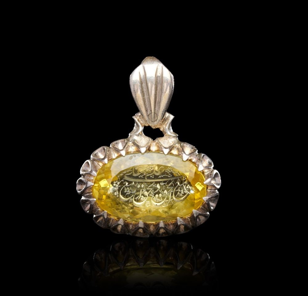 A CALLIGRAPHIC INSCRIBED AGATE AMULET ON SILVER, 19TH CENTURY, PERSIA: A CALLIGRAPHIC INSCRIBED YELLOW AGATE AMULET ON SILVER, PERSIA, 19TH CENTURY Of horizontal oval form, the faceted yellow agate set within a silver mount and surmounted by a fluted, bud-shaped suspensi