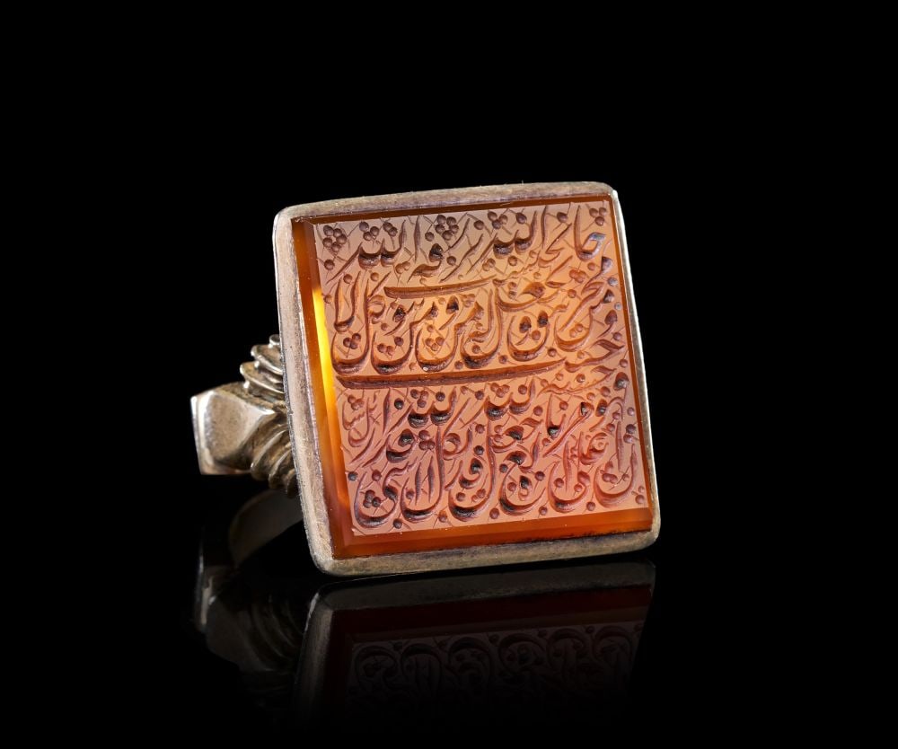 A CALLIGRAPHIC INSCRIBED AGATE RING ON SILVER, 19TH CENTURY, PERSIA: A CALLIGRAPHIC INSCRIBED AGATE RING ON SILVER, PERSIA, 19TH CENTURY The ring set with a square bezel of orange-toned agate, the stone polished flat and held within a plain silver collet raised above a