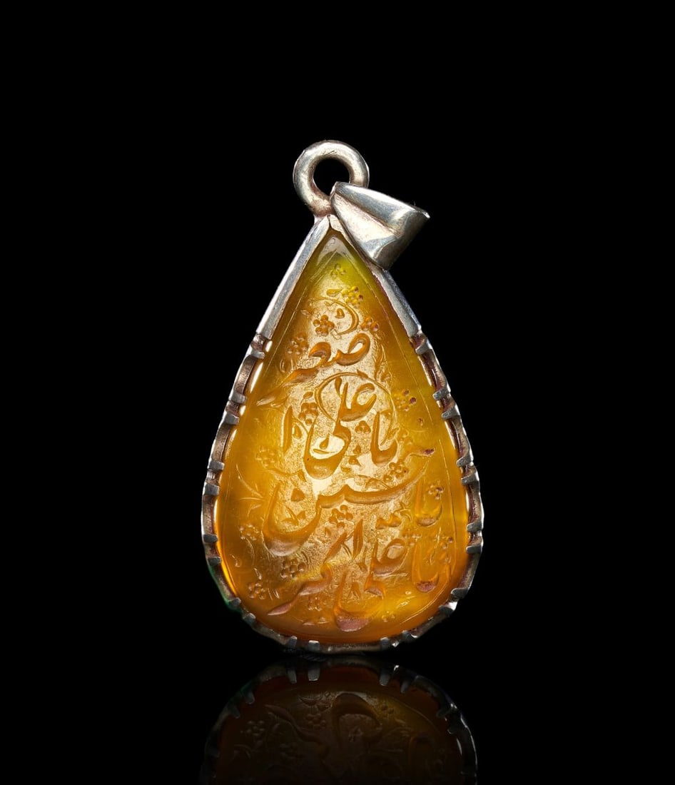 A CALLIGRAPHIC INSCRIBED AGATE AMULET ON SILVER, 19TH CENTURY, PERSIA: A CALLIGRAPHIC INSCRIBED AGATE AMULET ON SILVER, PERSIA, 19TH CENTURY Of pear-form, the translucent honey-coloured agate set within a silver mount with a suspension loop at the apex and a serrated bez