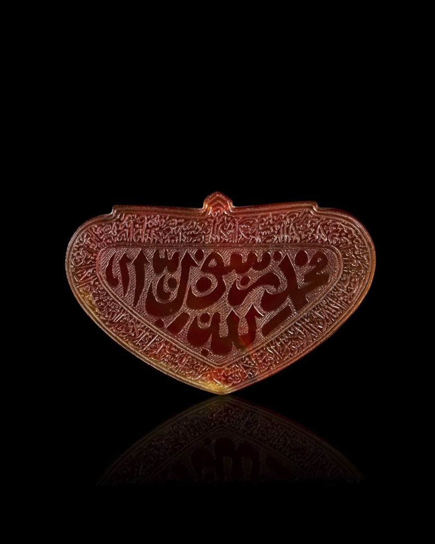 A CALLIGRAPHIC INSCRIBED AGATE SEAL, 19TH CENTURY, PERSIA: A QAJAR CARNELIAN CALLIGRAPHIC AMULET, PERSIA, 19TH CENTURY Of horizontal heart-shaped or shield form, surmounted by a small projecting tab pierced for suspension, the deep reddish-brown stone carved 