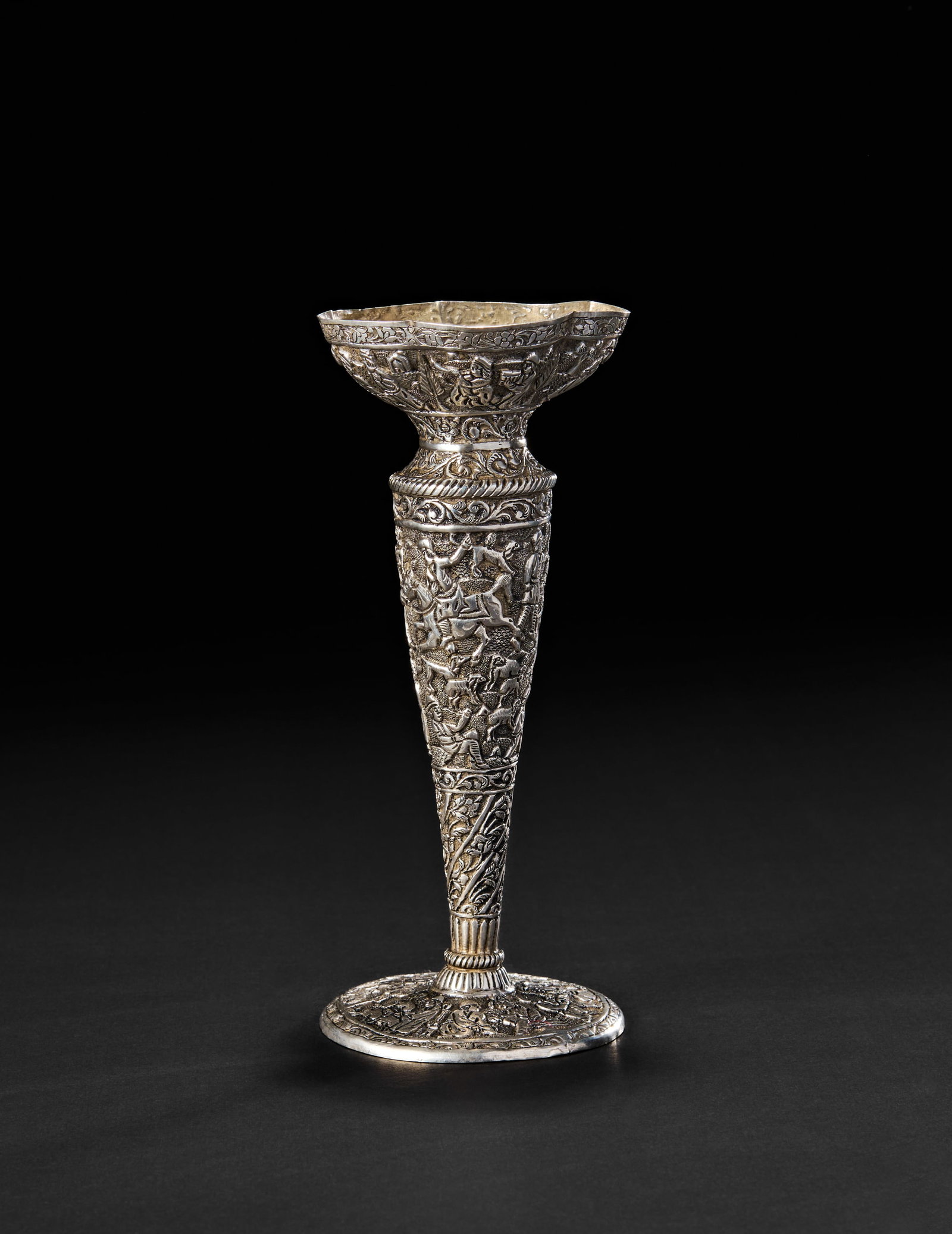 A PERSIAN SILVER TRUMPET VASE, SHIRAZ: Of a flaring form, this finely hand-chased silver vase is richly adorned with a dynamic band of figures and animals in various animated scenes, including horseback riders, musicians, and pastoral moti