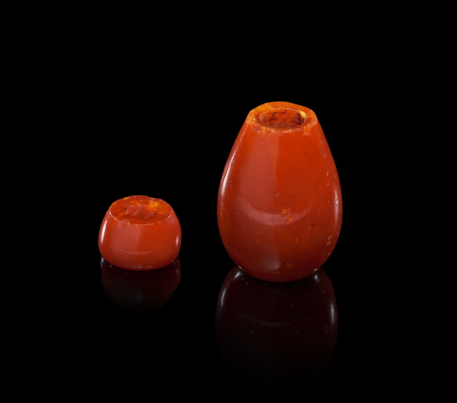 TWO OTTOMAN AMBER PIPE ORNAMENTS, TURKEY, 19TH CENTURY: Two Ottoman pipe ornaments carved from translucent orange-red amber. The collection includes a larger pear-shaped ornament and a smaller flattened discoid ornament, both featuring central drill