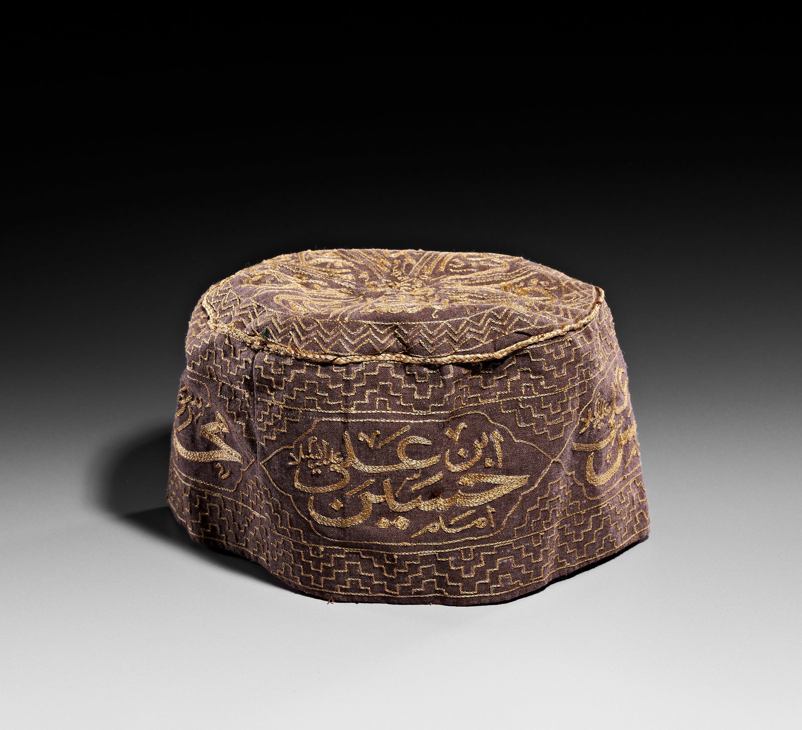 AN EMBROIDERED DERVISH HAT WITH CALLIGRAPHIC CARTOUCHES, 19TH CENTURY: This embroidered textile skullcap, identified as a dervish hat, is composed of a dark brown base fabric embellished with gold-toned threadwork. The crown features a radial arrangement of calligraphic