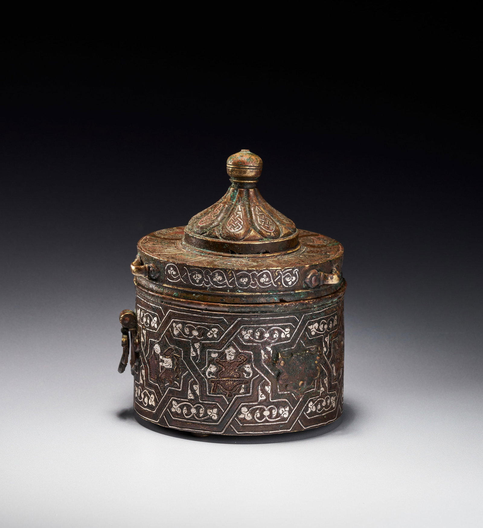 A SILVER-INLAID SELJUK COPPER ALLOY INKWELL, KHURASAN, 13TH CENTURY: This cylindrical lidded vessel is a silver-inlaid Seljuk inkwell, attributed to the Khurasan region, likely Herat, dating to the 13th century. The object is constructed from a copper alloy and feature