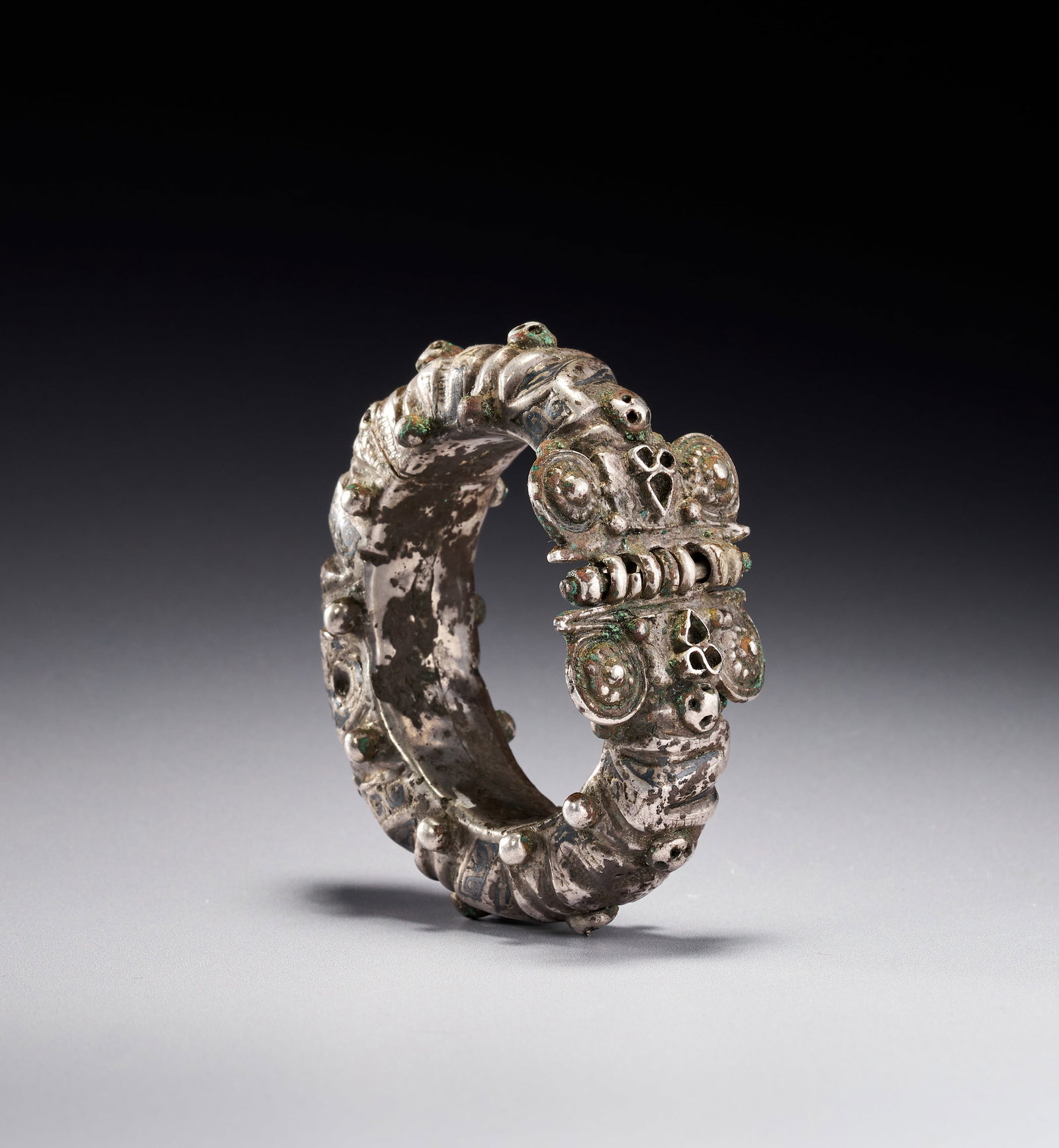 A SELJUK SILVER AND NIELLO CUFF BRACELET, IRAN, 12TH CENTURY: Seljuk silver and niello hinged bangle bracelet, attributed to 12th century Persia. The heavy, wide band features elaborate repousse and cast ornamentation. The exterior is divided into sections by fl