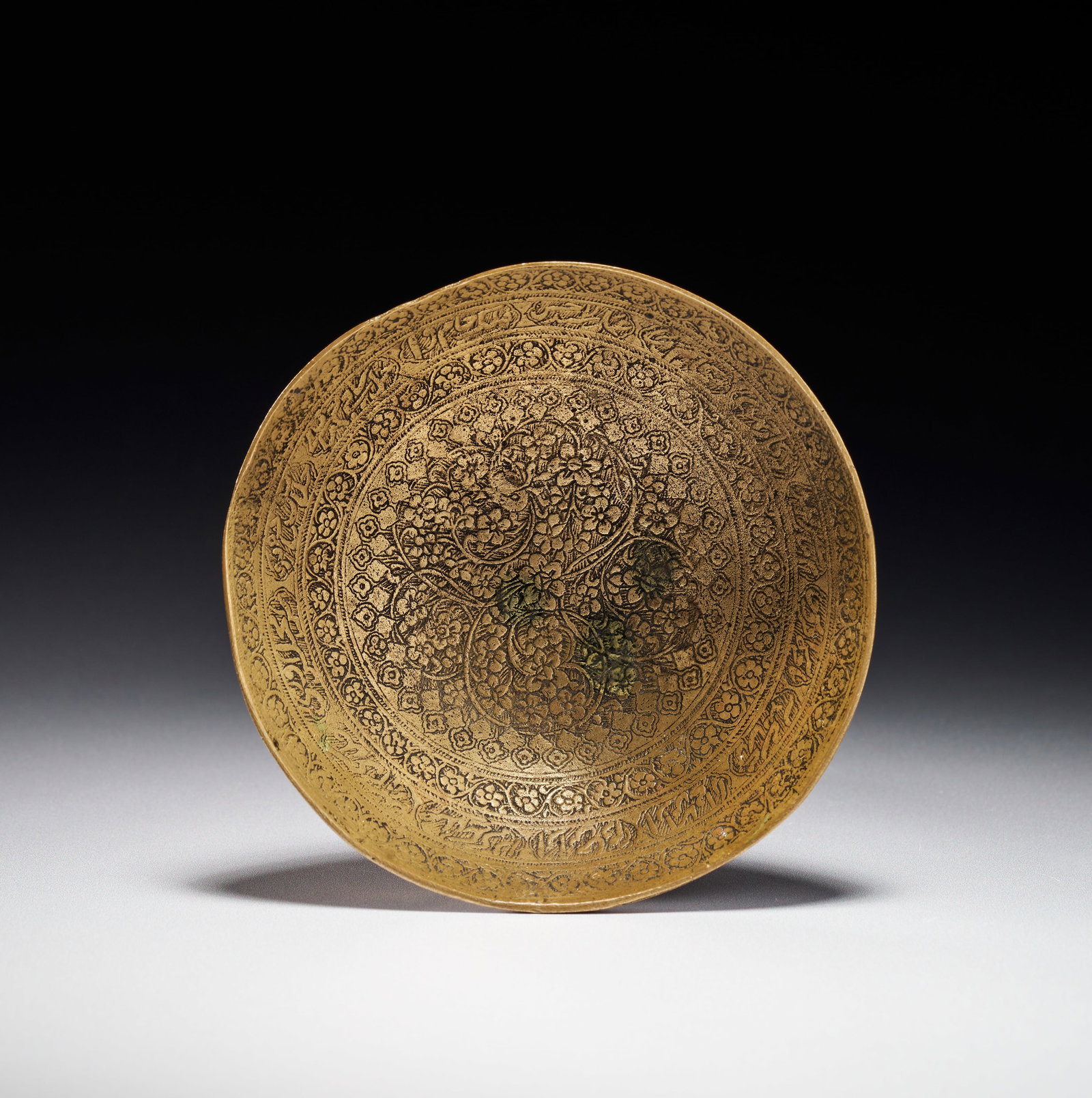 A PERSIAN ZAND ENGRAVED BRASS DRINKING BOWL WITH CALLIGRAPHIC SCRIPT, 18TH CENTURY: Features a plain, rounded exterior and a densely decorated interior. The interior surface is executed with fine engraving and etching, centered on a large medallion containing floral and scrolling vin