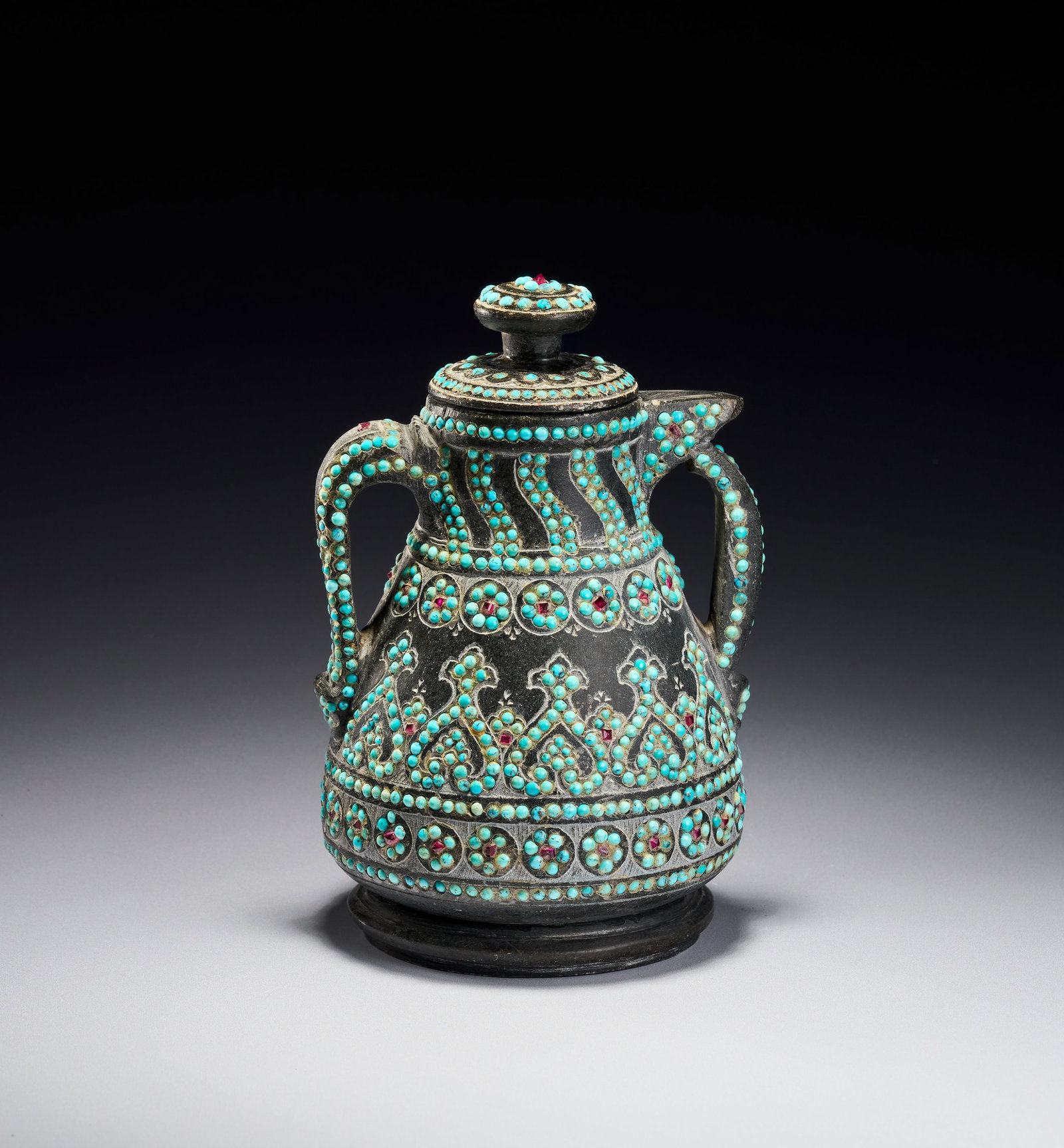 A RARE QAJAR TURQUOISE INSET CARED STONE TEAPOT, PERSIA, 19TH CENTURY: The vessel is carved from a dark hardstone, such as chlorite or soapstone, and features a matte patina. The body is heavily ornamented with inset gemstone cabochons, including turquoise and small red