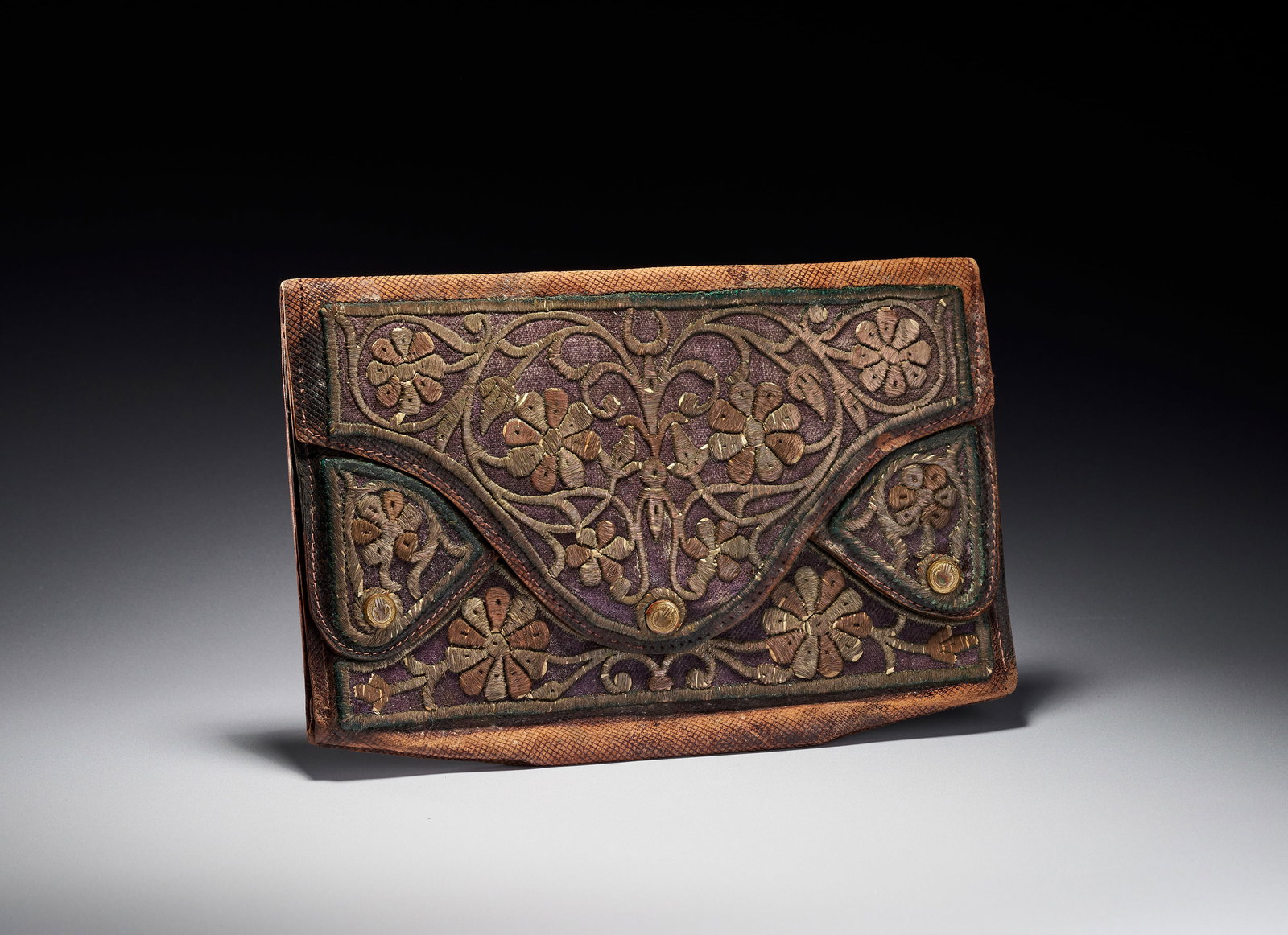 AN OTTOMAN GILT-THREAD EMBROIDERED LEATHER WALLET, TURKEY, 18TH CENTURY: The exterior is constructed from dark brown leather with a fine cross-hatched grain. The front features inset purple fabric panels heavily embellished with floral and foliate embroidery in gold and co