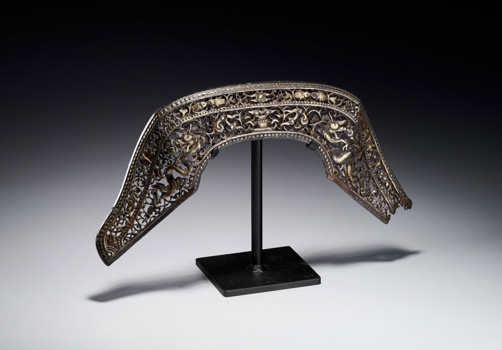 A CHINESE OR SINO-TIBETAN PIERCED IRON AND PARCEL-GILT SADDLE CANTLE PLAQUE, 16TH - 17TH CENTURY: This object is a decorative iron saddle fitting, identified as a cantle plaque, featuring an arched form with downward-sloping lateral wings. The piece is constructed from wrought iron using an intric
