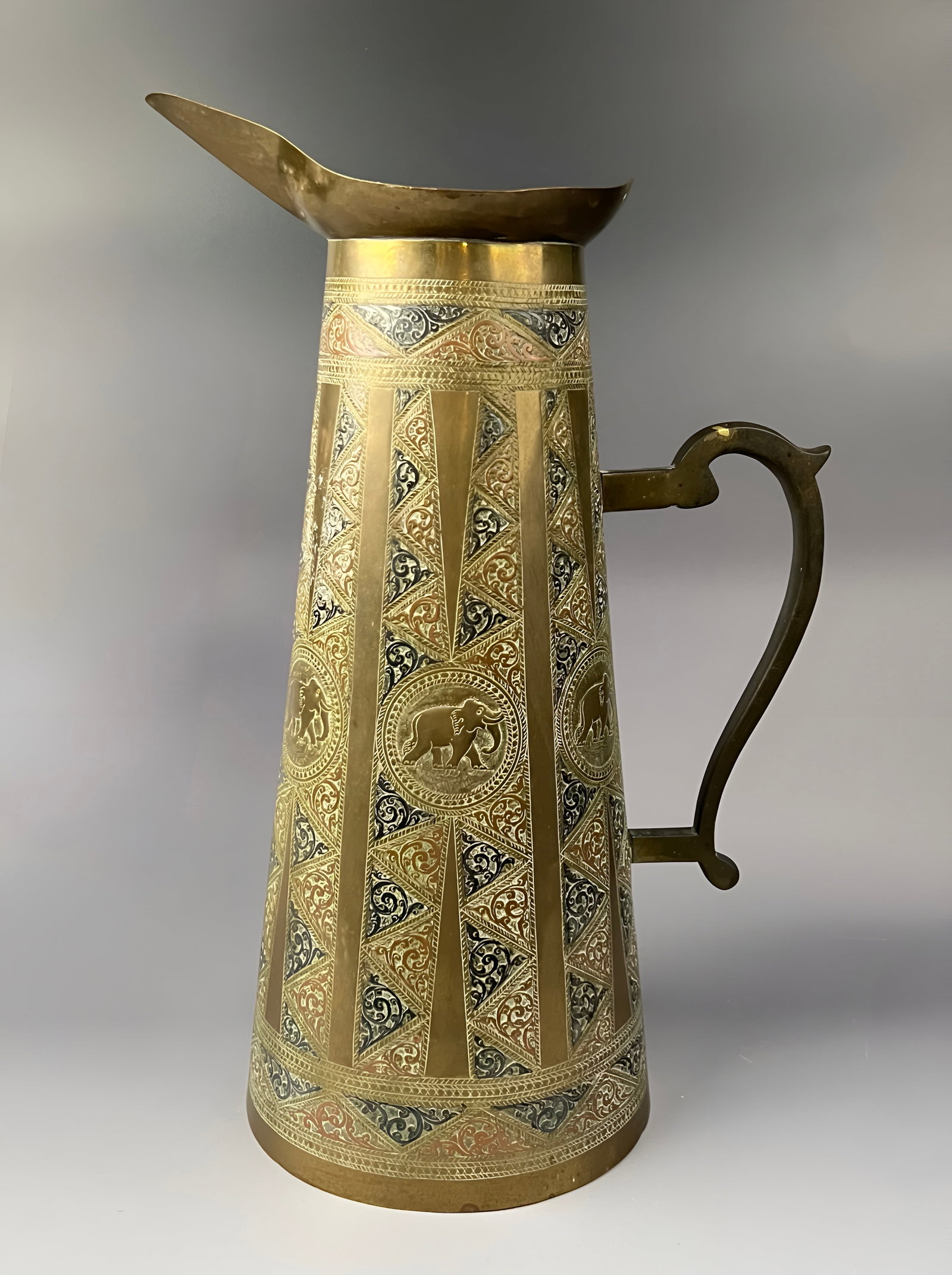 A SRI LANKAN MIXED METAL INLAID BRASS EWER WITH ELEPHANT MOTIFS, 19TH CENTURY: A tall, tapered brass ewer featuring tri-colour mixed-metal inlay of copper and white metal. The exterior body is decorated with vertical panels of fine-line chased foliate and arabesque scrolling pat