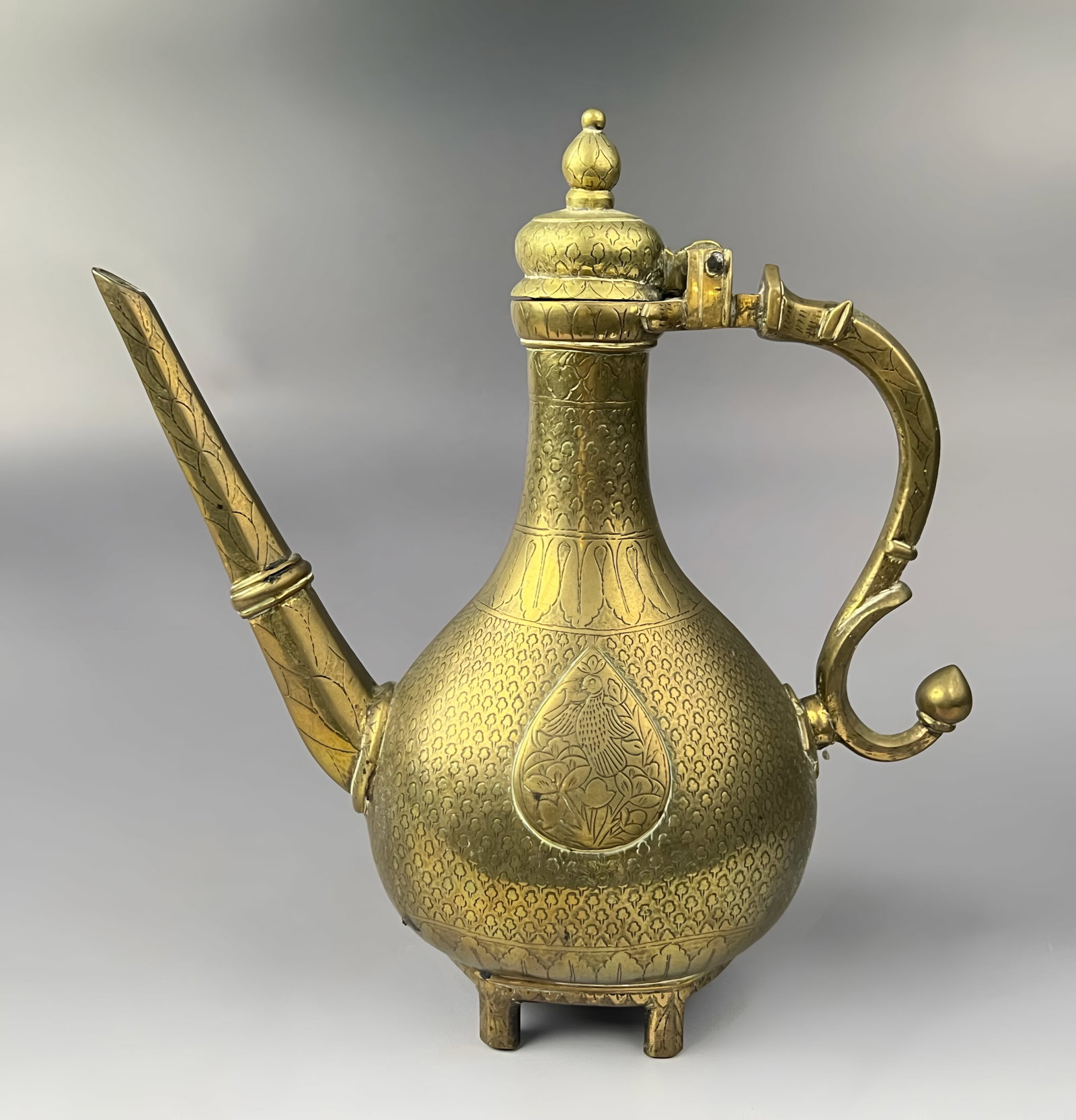A NORTH INDIAN BRASS AFTABA EWER WITH CHASED BIRD AND FLORAL DECORATION, 19TH CENTURY: An Indo-Persian brass ewer, known as an aftaba, featuring a pear-shaped body supported by three short feet. The vessel includes a long, straight, tapered spout and a scrolling C-shaped handle with a b