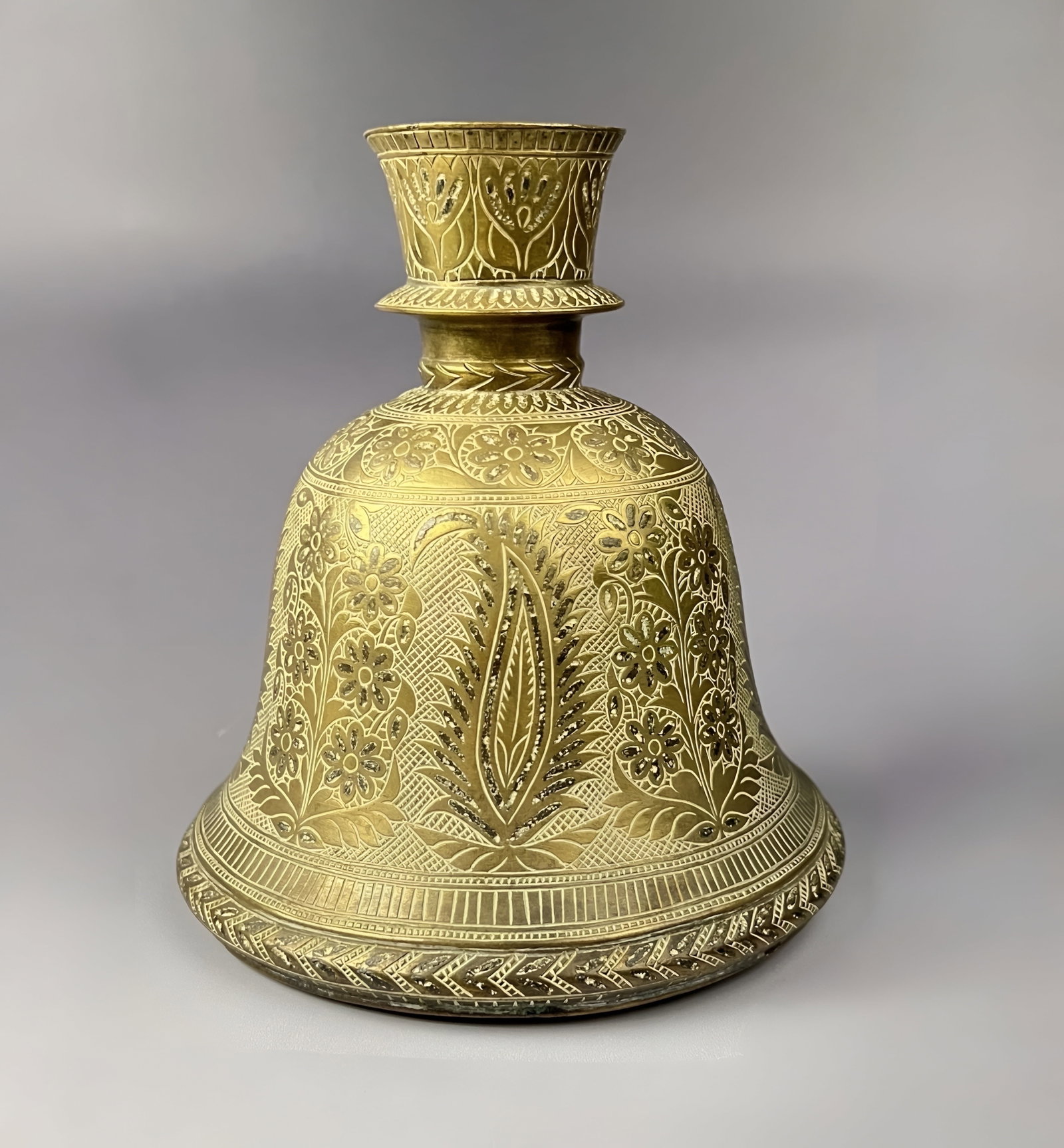 A MUGHAL BRASS HOOKAH BASE, LAHORE, EARLY 18TH CENTURY: The piece features a short, waisted neck that terminates in a flared, trumpet-shaped mouth. The exterior surface is entirely decorated with elaborate chased and engraved motifs. The central body is ad