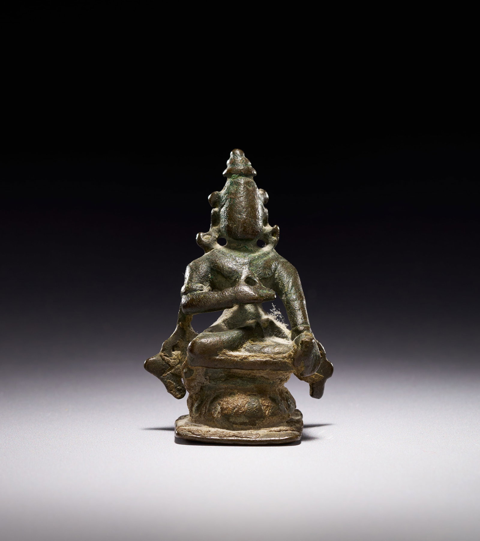 A SOUTHEAST INDIAN CAST BRONZE FIGURE OF A SEATED DEITY, 18TH CENTURY: The figure is adorned with a tall, conical jatamakuta crown, large circular earrings, and simple body jewellery. The right hand is raised in a mudra, while the left hand rests in the lap. Two stylised