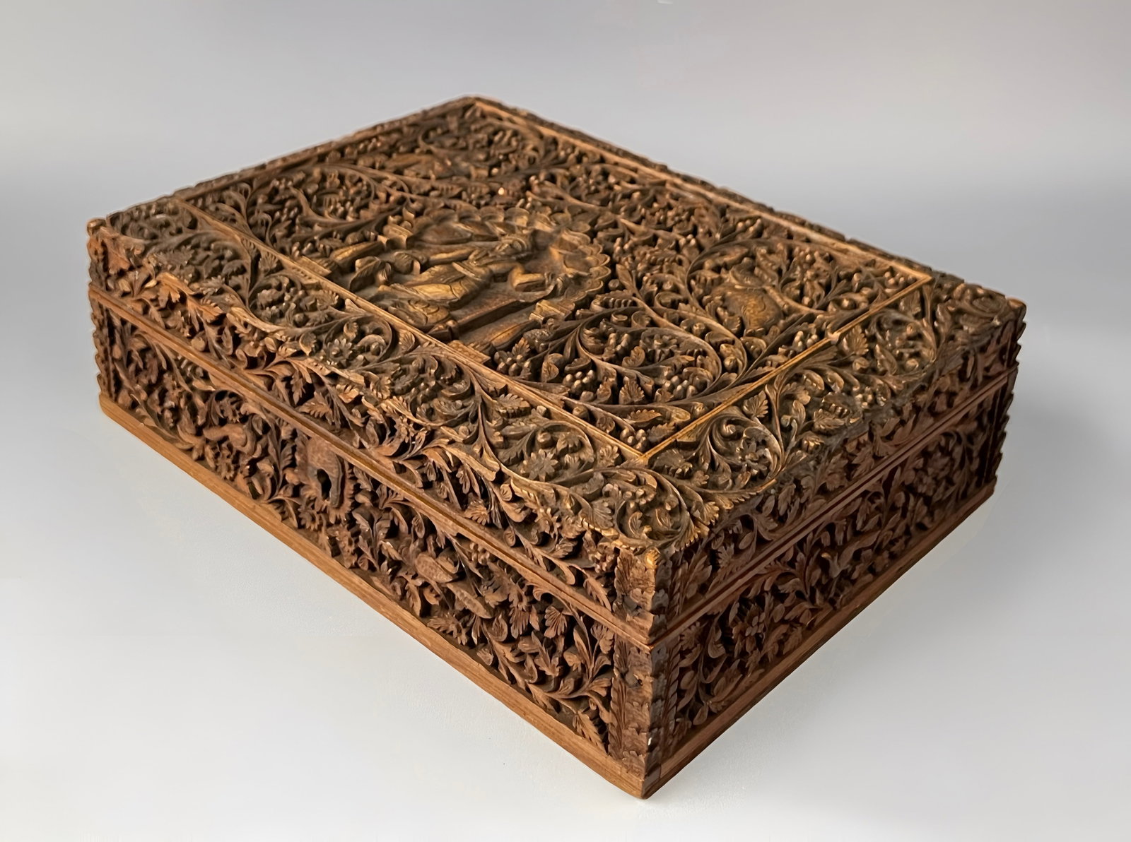 A MYSORE CARVED SANDALWOOD JEWELLERY BOX, INDIA, LATE 19TH CENTURY: A rectangular box constructed from hand-carved sandalwood, featuring exceptionally deep high-relief decoration across all exterior surfaces. The lid is centred by a rectangular panel depicting a seate
