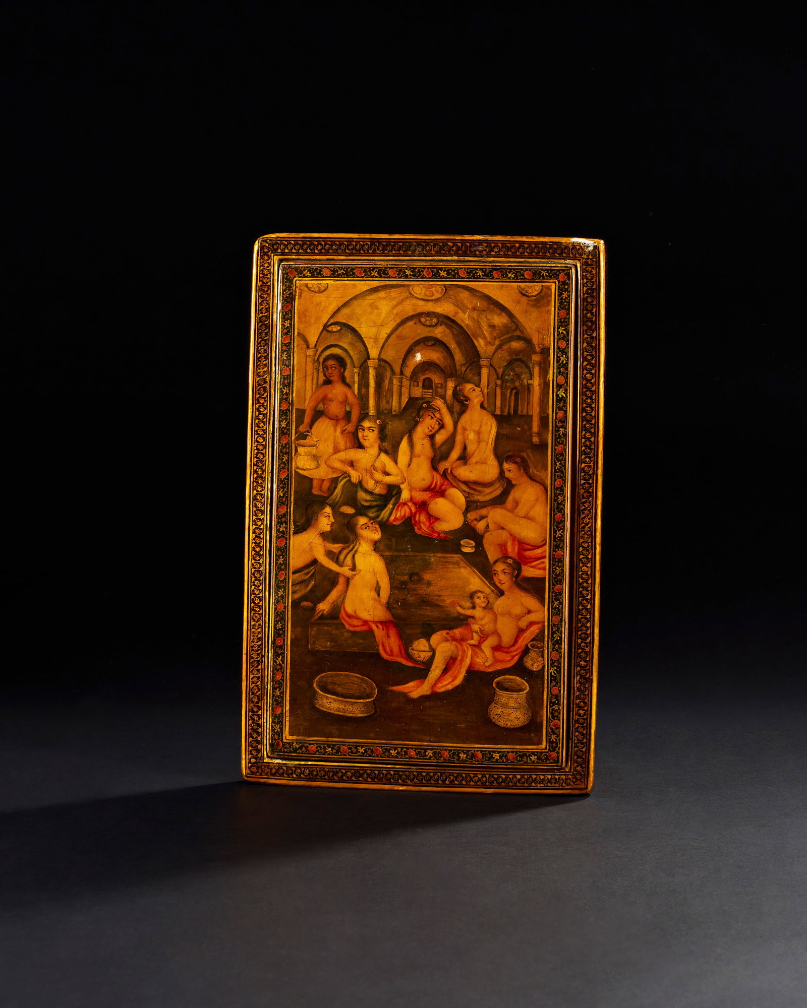 A 19TH CENTURY QAJAR LACQUERED MIRROR CASE SIGNED YA SAHIB AL-ZAMAN AND DATED: A 19th century Qajar lacquered mirror case portrays a vibrant and detailed scene of a ladiesâ€™ hammam (bathhouse). The front panel is adorned with an intricate painting of several women, eleg