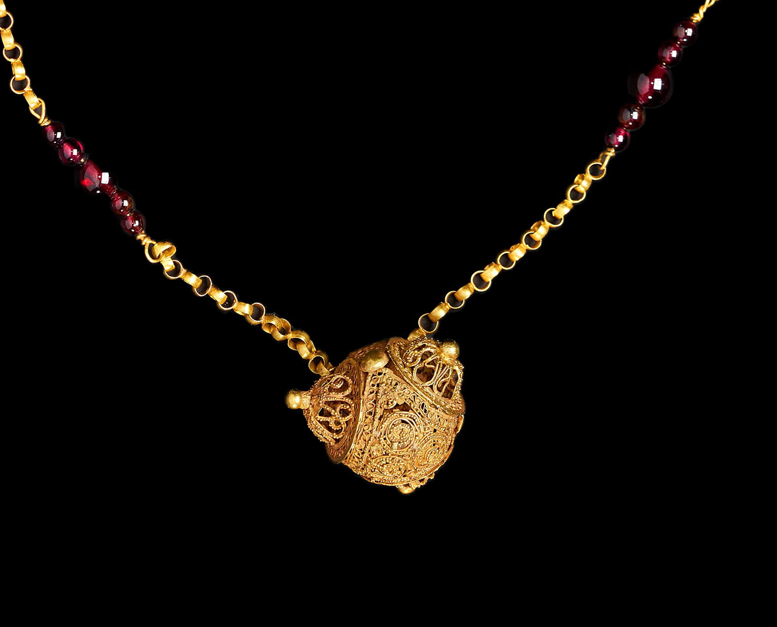 A GOLD FATIMID FILIGREE GOLD PENDANT NECKLACE WITH GARNET BEADS, EGYPT OR SYRIA, 10THâ€“12TH: A necklace featuring a central spherical pendant decorated with detailed filigree wirework and granulation. The pendant is constructed from a yellow metal and features several small raised domes and c