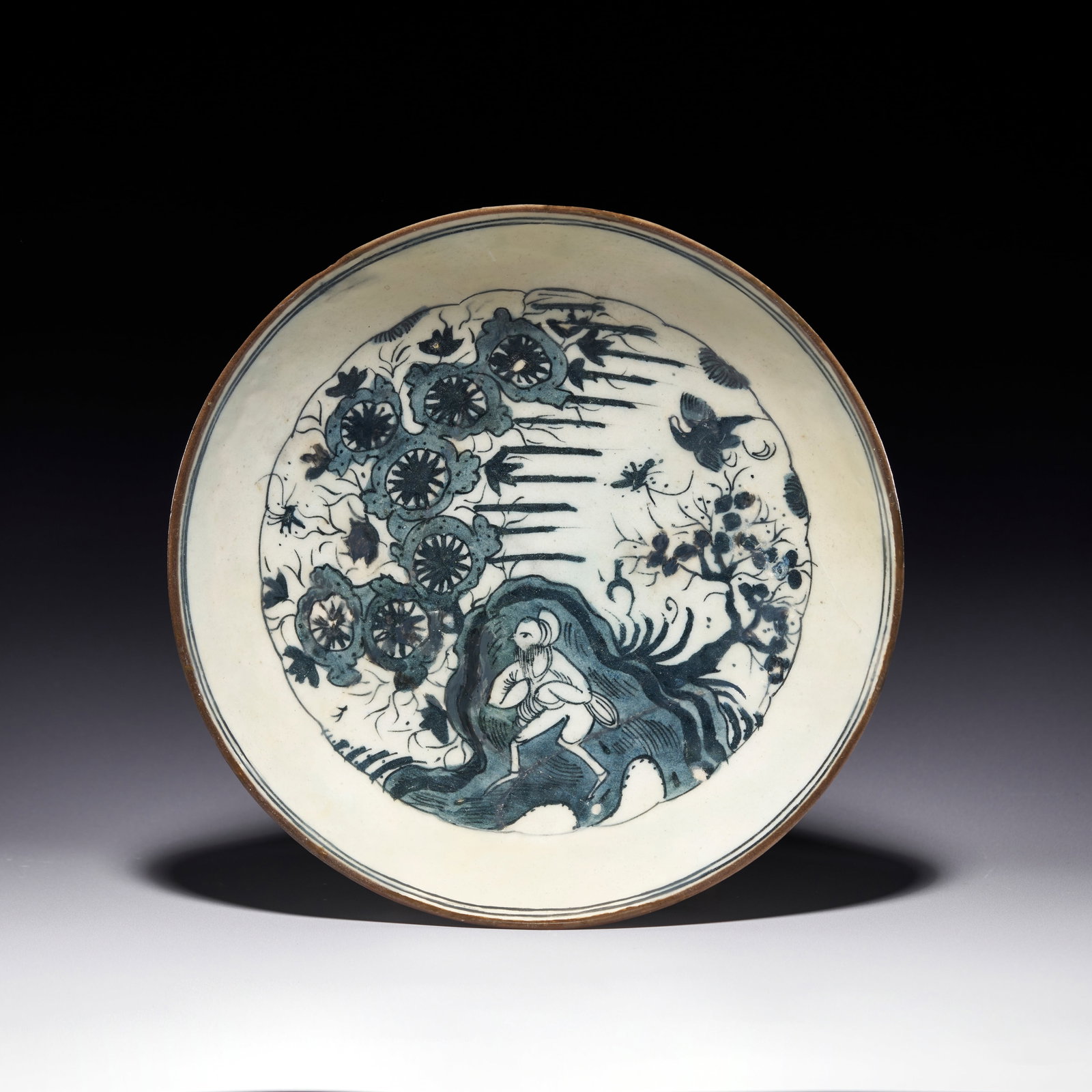 A BLUE AND WHITE CHINESE MOTIFS SAFAVID CERAMIC DISH, PERSIA, 17TH CENTURY: The dish is of shallow, rounded form with a recessed footring and is decorated in underglaze blue beneath a light celadon-tinted glaze. The interior depicts a central figure, possibly a scholar or an 