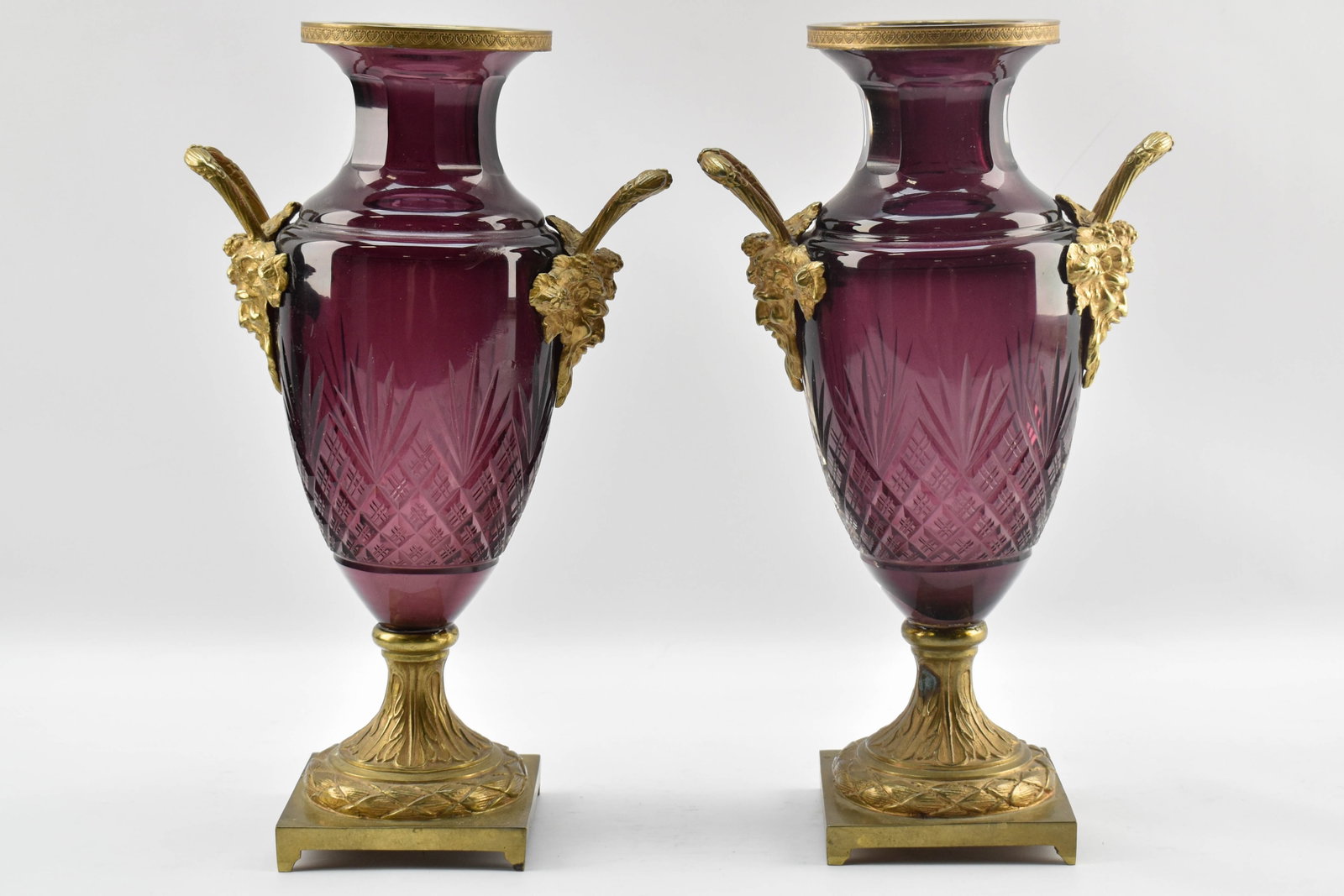 Pair of Vintage Purple Vases: Pair of pedestaled vases with wine-colored, diamond-cut glass bodies and gold-toned rims, handles, and bases, possibly bronze. The handles are shaped like the face of the Greek god Dionysus and includ