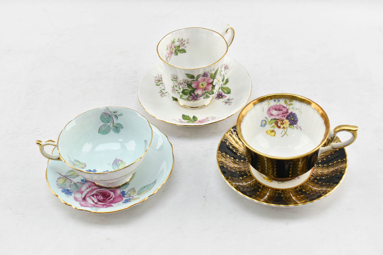 Three Paragon China Teacup and Saucer Sets: Three vintage teacups and matching saucers made of fine bone china by the British Paragon China Company (1919-1960). Includes a floating rose on black and gold set circa 1957, a pink cabbage rose on m