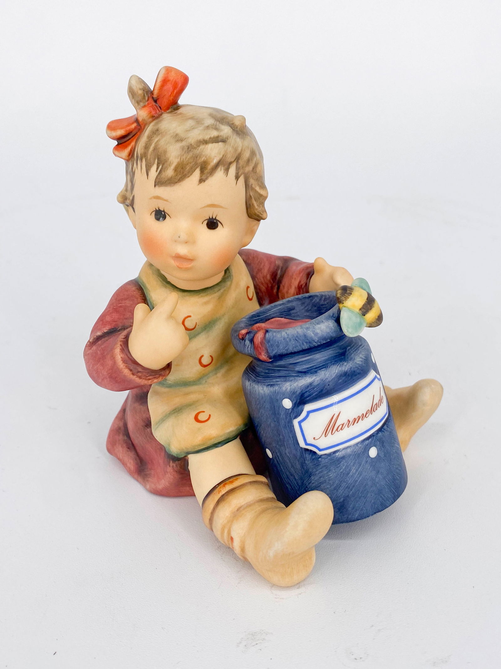 Goebel Hummel Marmalade Lover Figurine 2249: Ceramic figurine by Goebel Hummel titled Marmalade Lover depicting a little girl rwith a large jar of marmalade in her lap. Marked on underside with Goebel logo. Sticker adhered to back and signed on