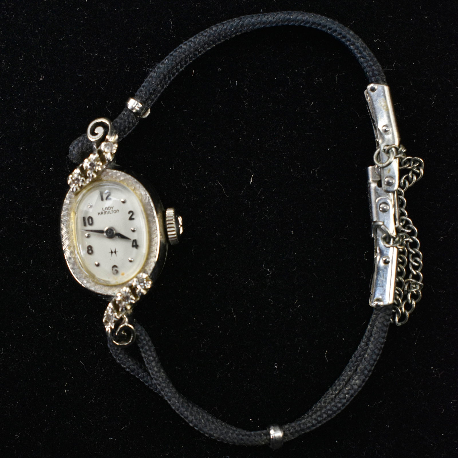 White Gold Lady Hamiliton Wrist Watch: A white gold Lady Hamilton wrist watch with a black cord band, stamped on case back “Hamilton 14K gold”. The face of the watch is stamped “Lady Hamilton” and the lugs of the wa