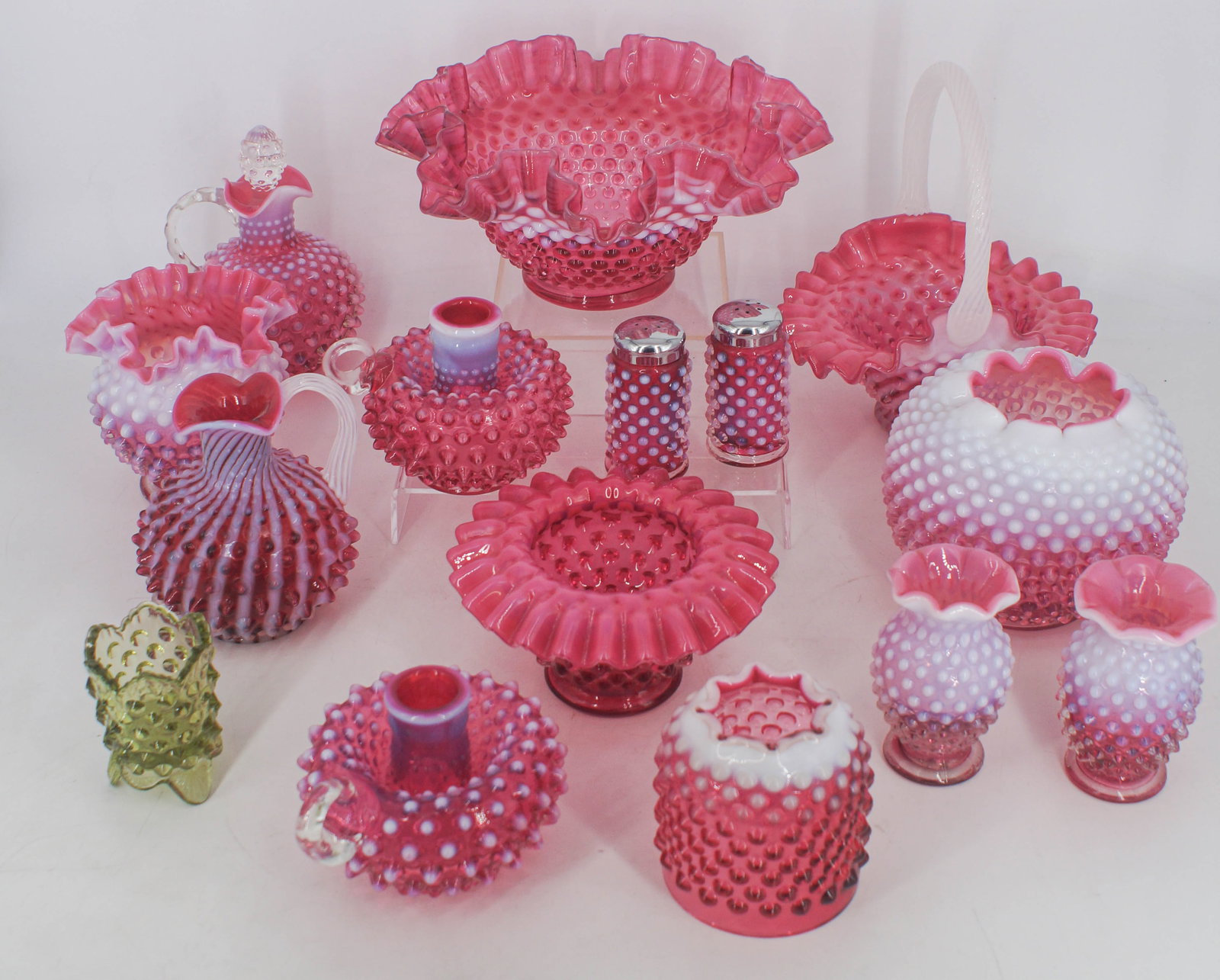 15 Assorted Cranberry Glass Hobnail Vessels: Assorted lot of hobnail glass, all cranberry with the exception of one green vessel. Lot includes a salt and pepper shaker, a handled basket, 2 pitchers/creamers, 2 candlestick holders, and more, as p