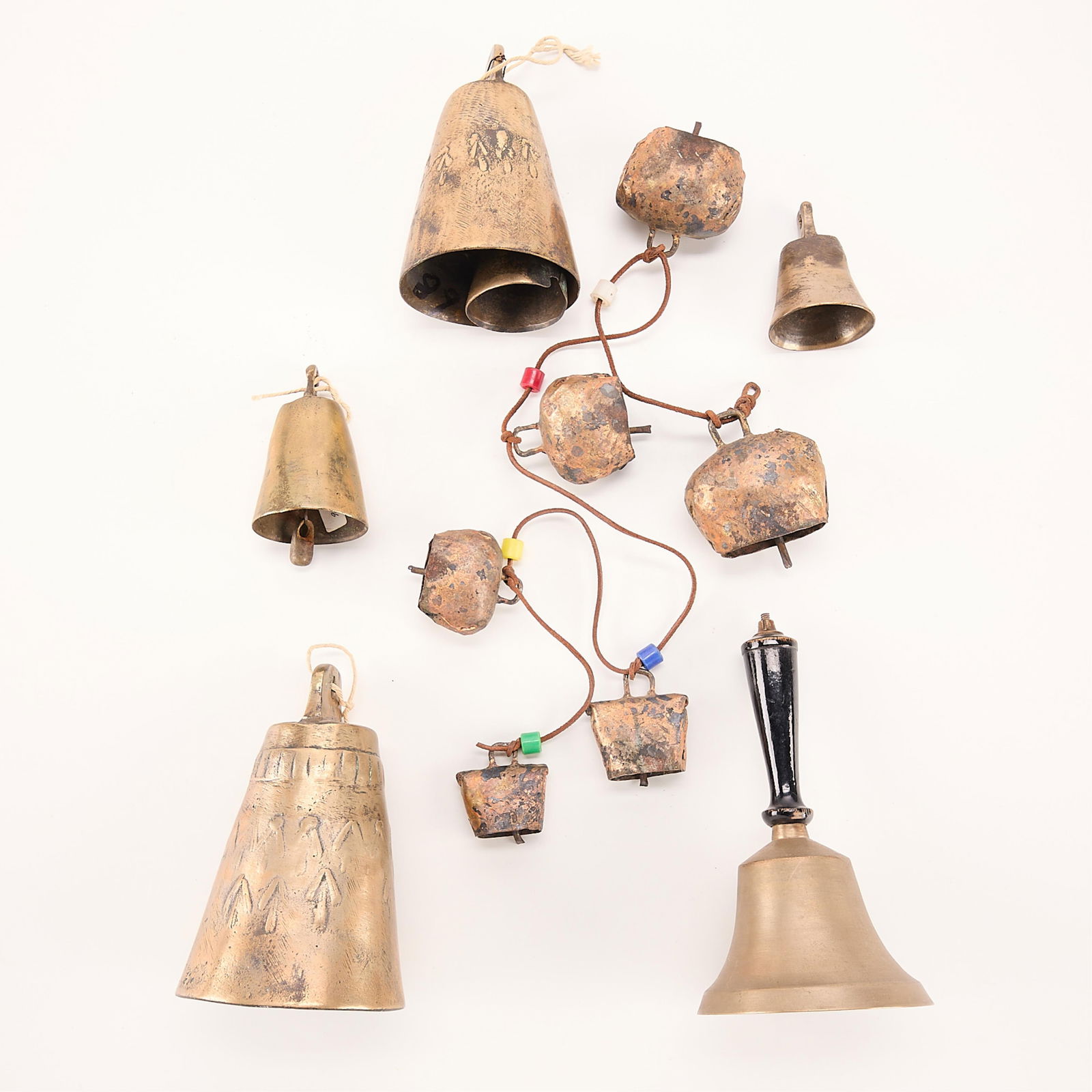 6 Metal Sheep Bells: Six metal sheep bells on brown leather string. All bells are varying sizes. Five colorful plastic beads sit in between all bells. Bell with another bell inside. Arrow decoration on outside. Small bras