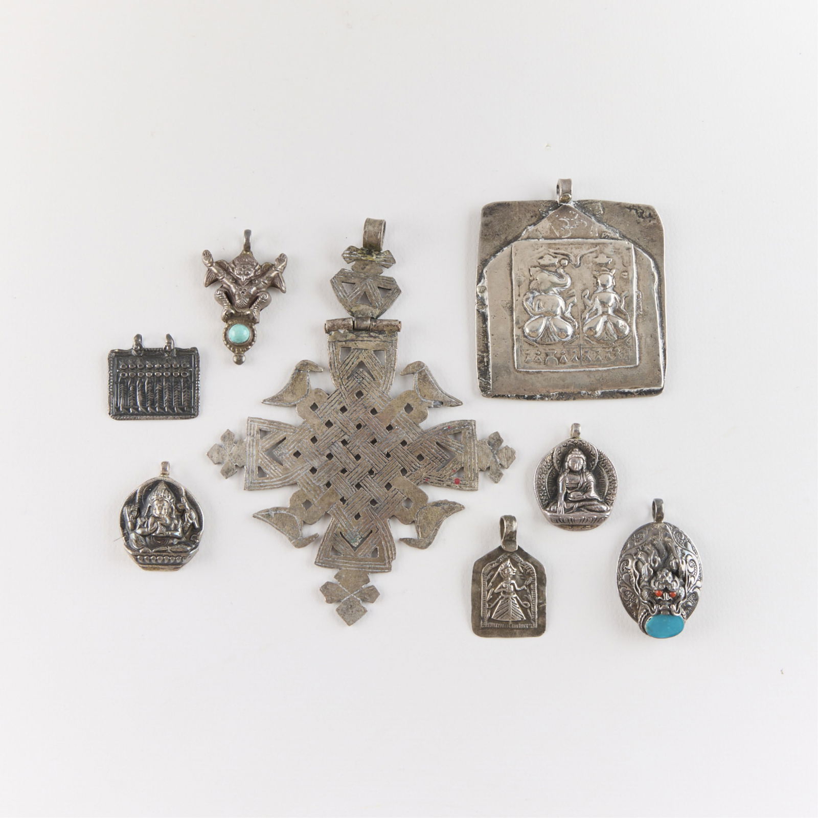 8 Asian, Southeast Asian, & Ethiopian Pendants: Including one pendant depicting the Ethiopian Coptic cross, five Indian pendants or amulets depicting Hindu deities, one pendant depicting Buddha, and one dragon head pendant with inlaid stones.Gross