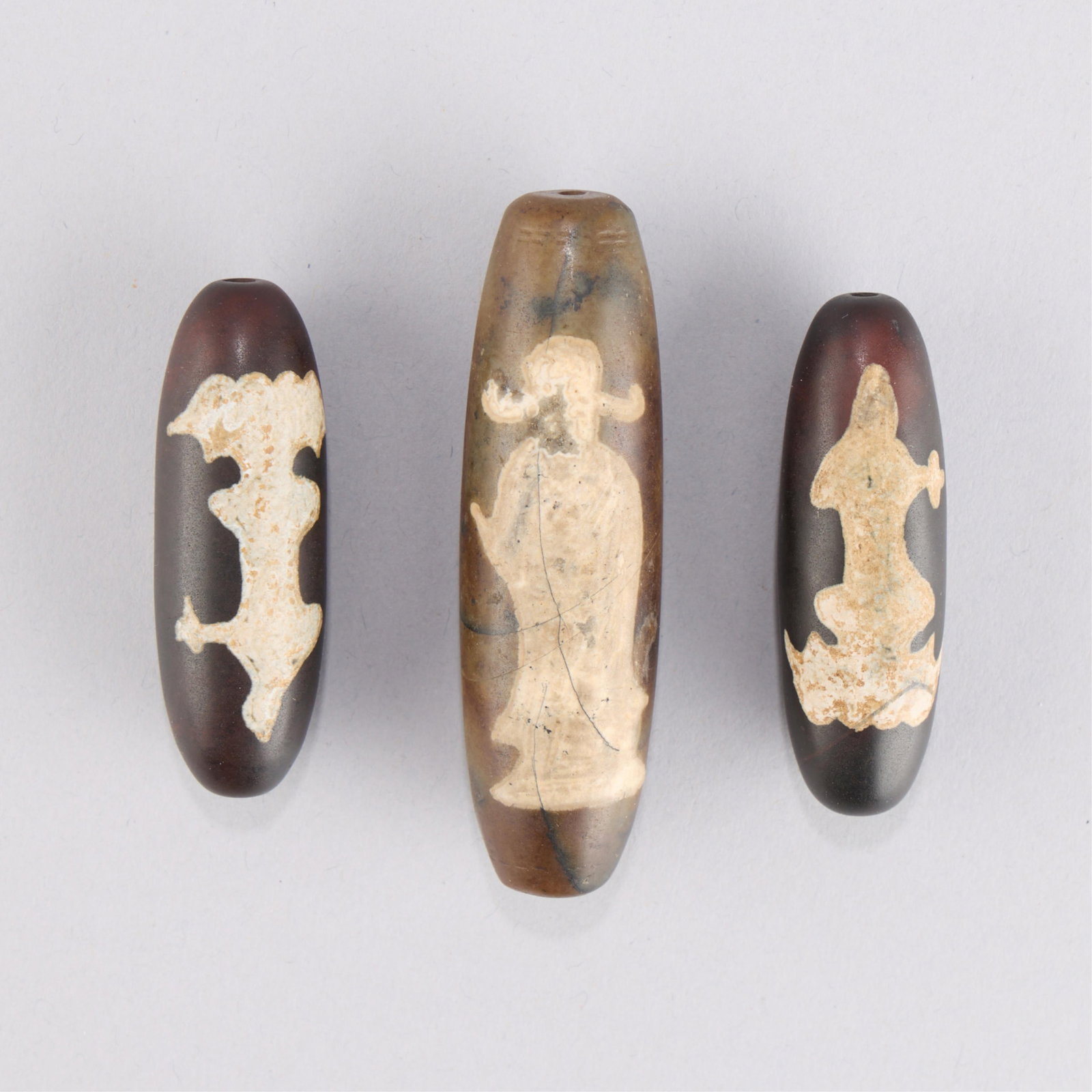 3 Tibetan Agate Dzi Beads: Group of three Tibetan agate dzi beads. Each with a symbol along one side and a guanyin form along the other. Length ranges from 2 1/8 in to 3 in; diameter ranges from 5/8 in to 7/8 in. Gross weight o