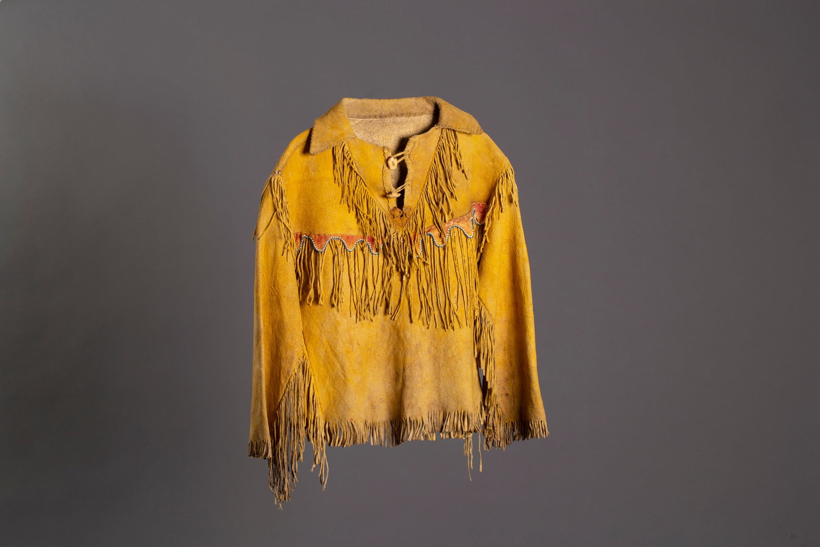 Southern Plains Child's Beaded Hide Shirt, Ca. 1900: Southern Plains Child's Beaded Hide Shirt Thread-sewn with a scalloped band coated in red pigment and edge-beaded in white and pony trader blue across chest and back, antler buttons, Ca. 1900. Length: