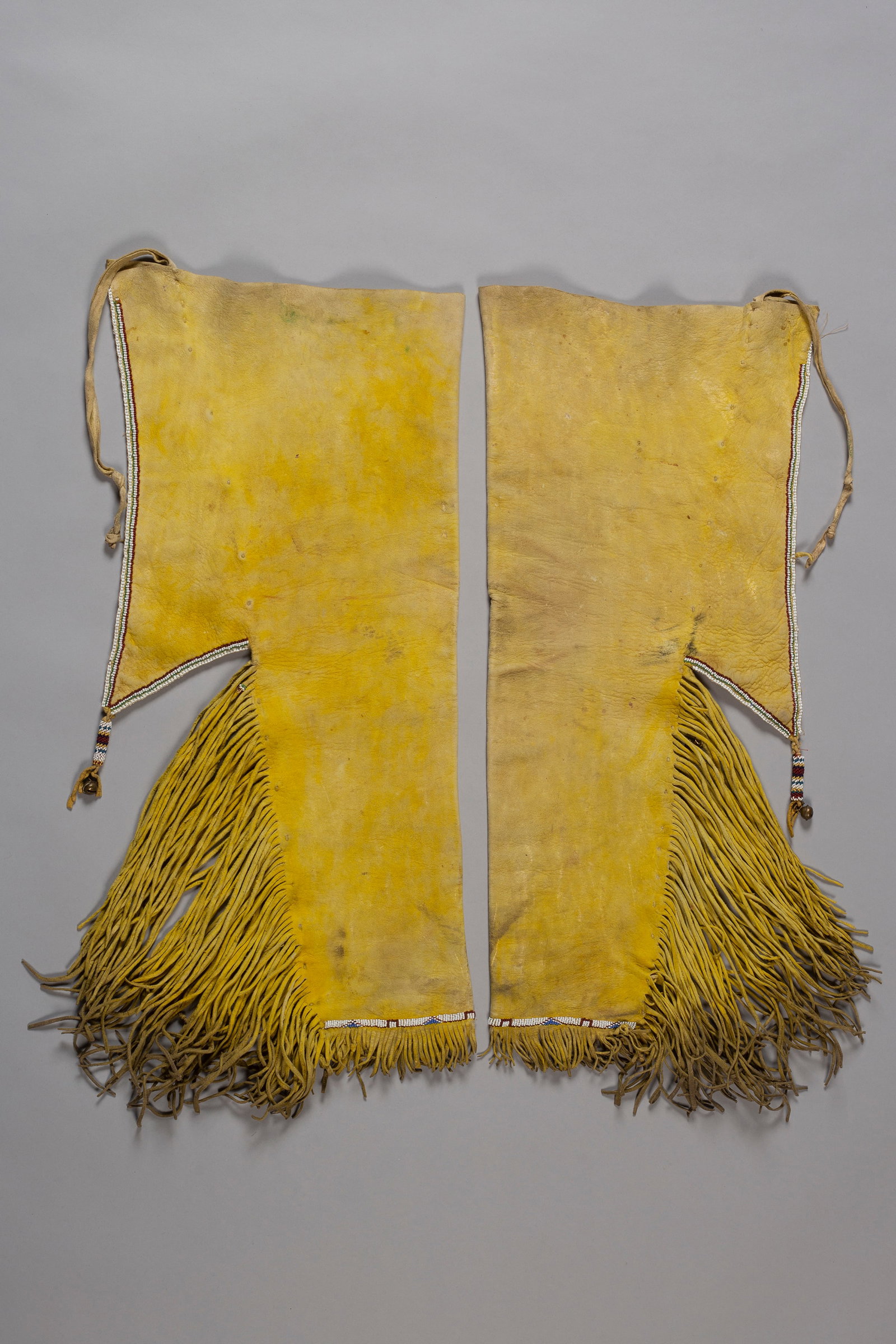 Kiowa Leggings, Ca. 1890s: Kiowa hide leggings with bead trim and beaded tassels with bells, Ca. 1890s. Fine leather fringe on lower half of each leg. Length with fringe: 26"