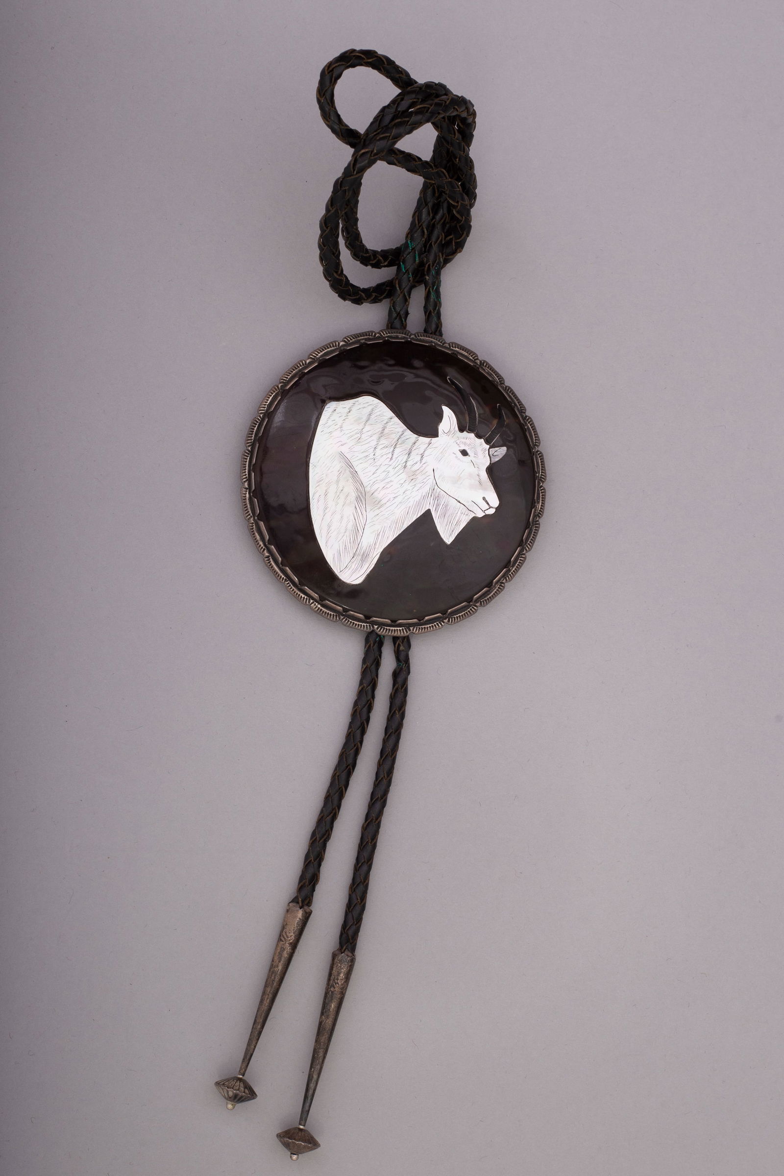 Elliot Qualo, Zuni Bolo Tie: White Mountain Goat on Black: Black leather bolo tie with round sterling silver clasp inlayed with a white mountain goat on black background by Elliot Qualo. Length: 39", Clasp diameter: 3 3/8", Weight: 82 grams; Provenance: Priva