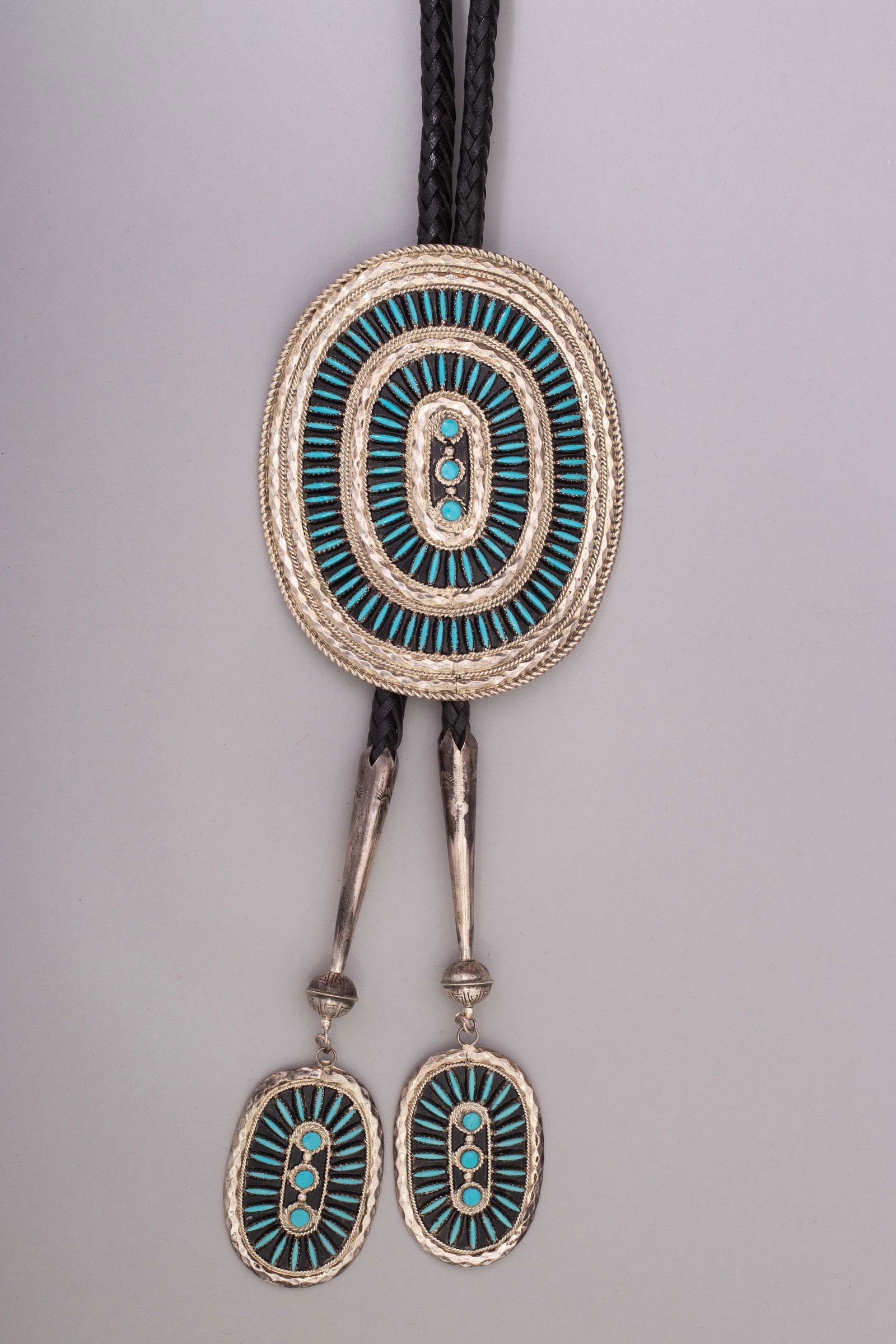 Zuni Needlepoint Bolo Tie: Black leather bolo tie with large silver and needlepoint turquoise clasp with silver cones capped with silver beads and matching silver and Zuni turquoise tips. Length: 54", Clasp: 4 1/2"x 3 3/4", Con